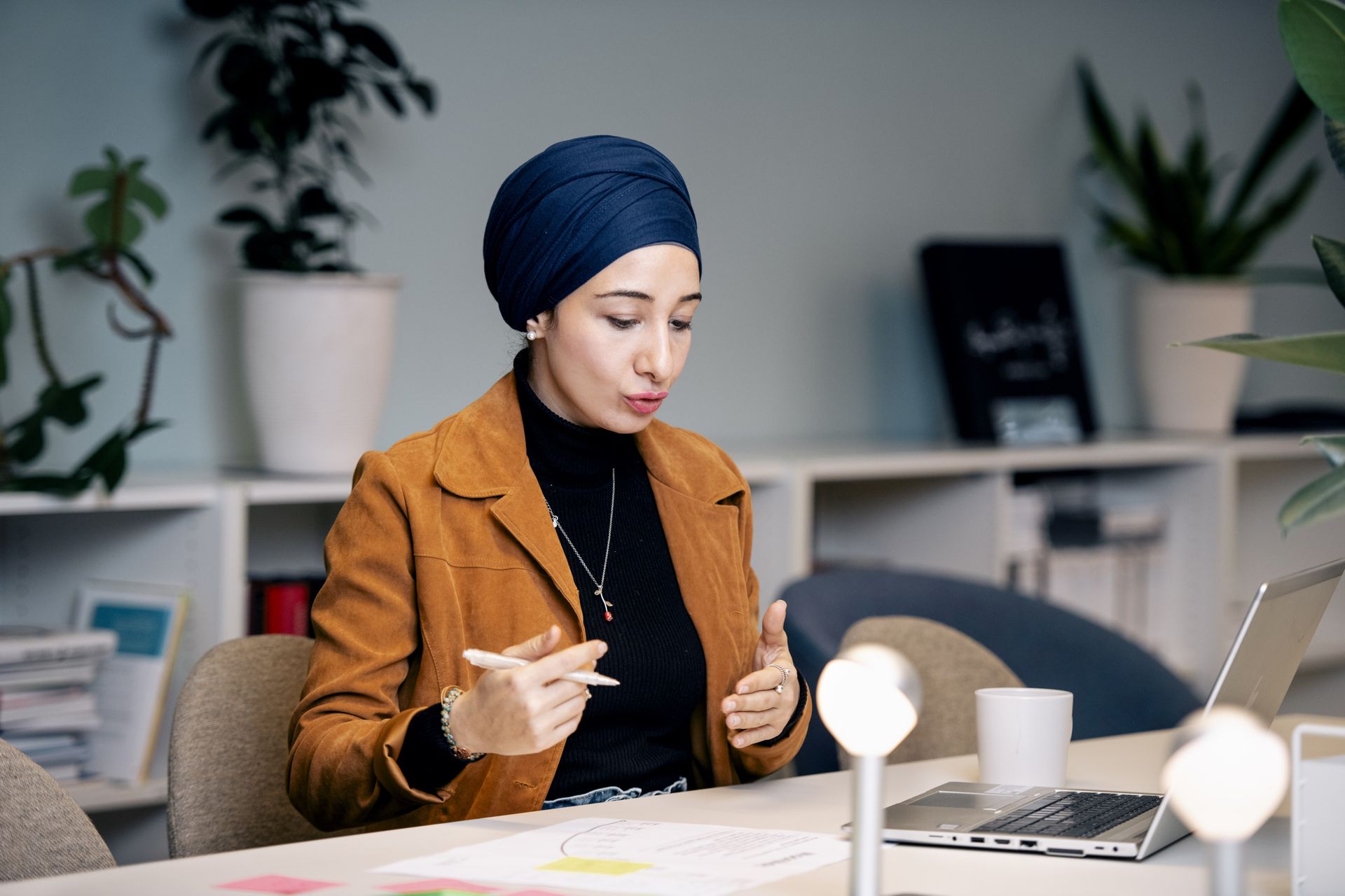 A woman sits by a desk, holding a pen. There's a laptop on the desk.