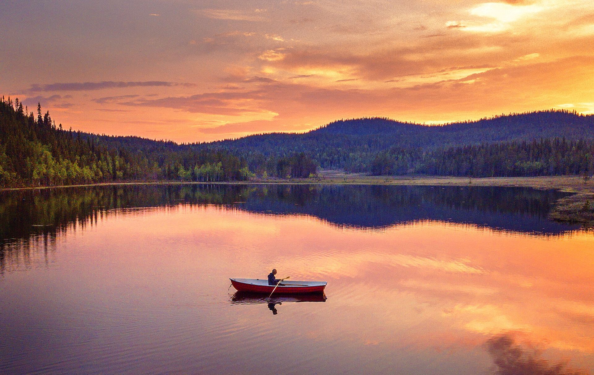 A person is rowing a boat on a mirrored lake under the midnight sun in Swedish Lapland.