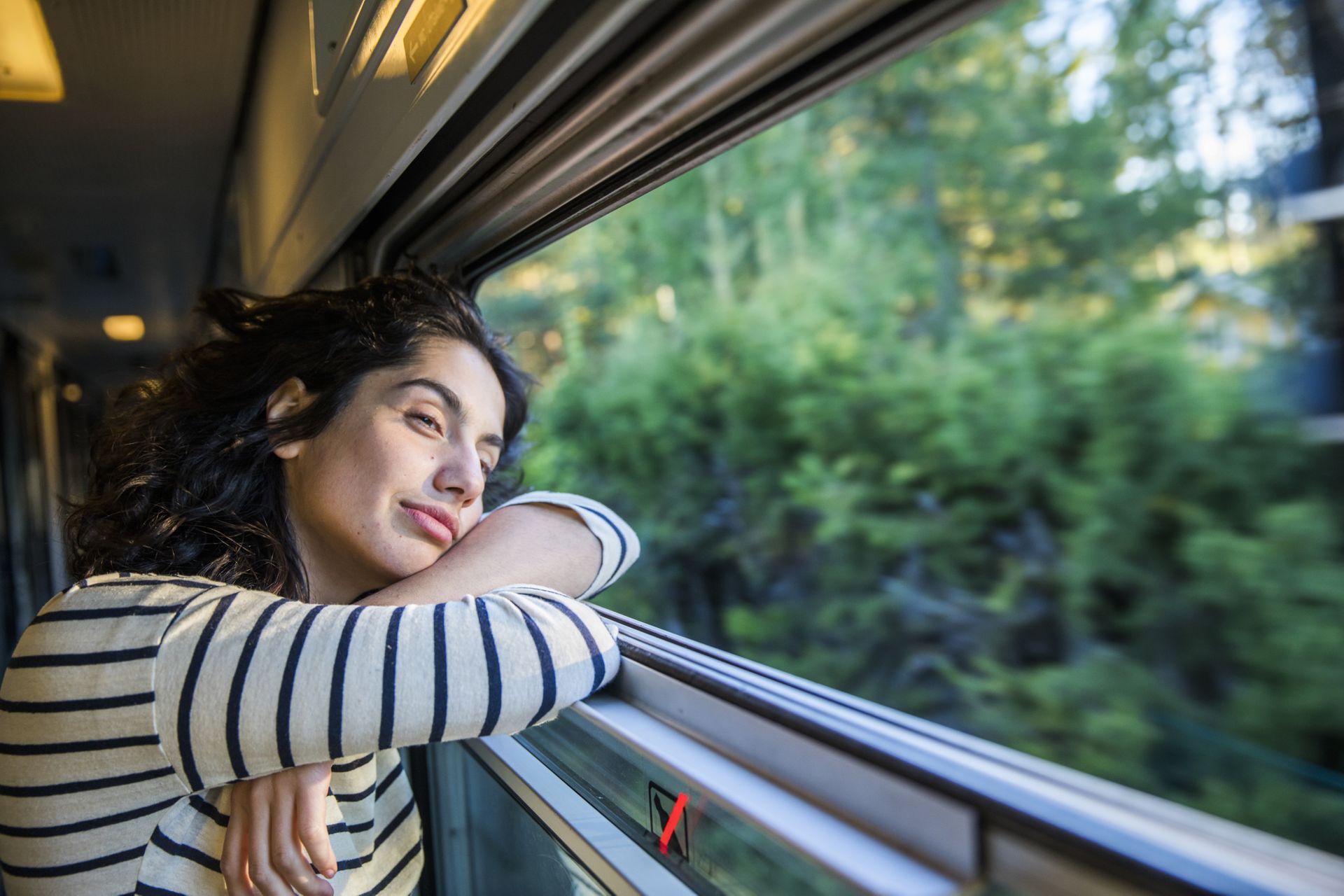 A woman is looking out of the window on a train while passing forest and greenery.