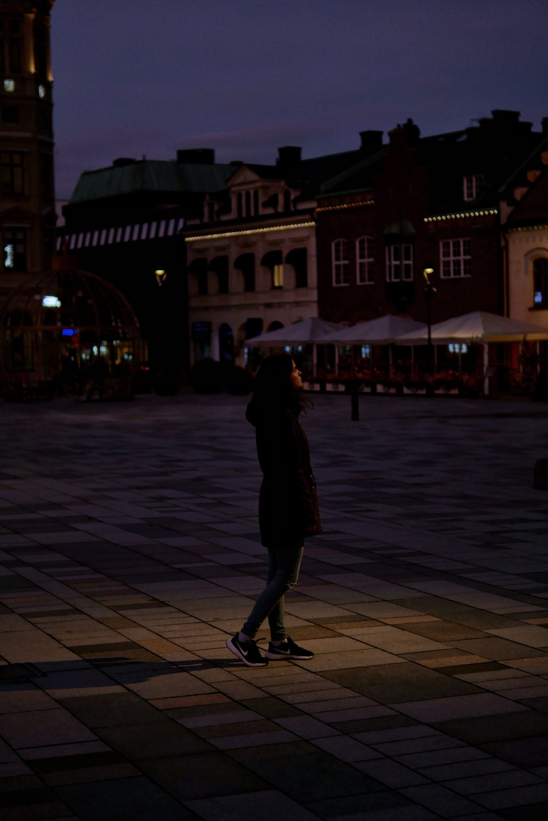 A woman illuminated by a spotlight, standing in a town square.