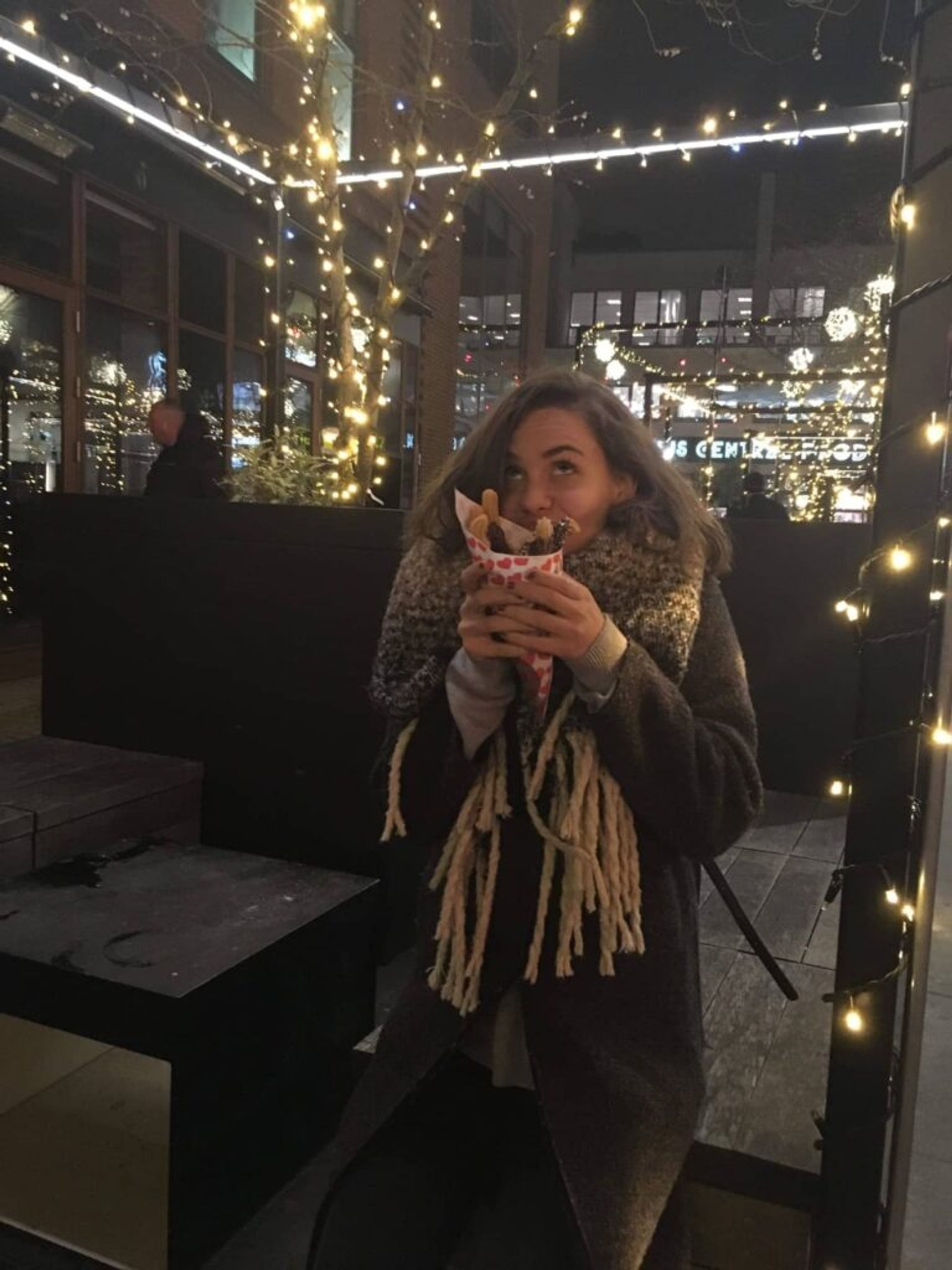 A woman savoring a treat outdoors, against a backdrop of festive lights.