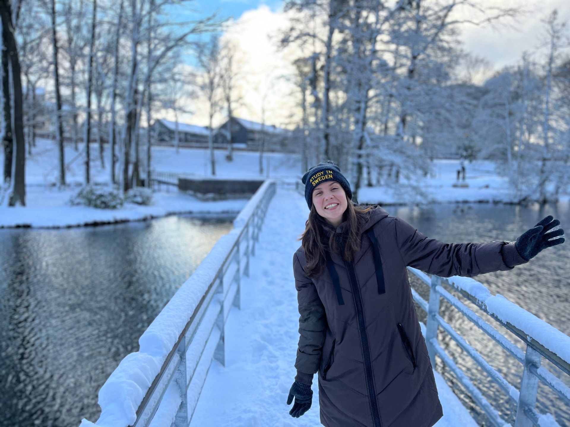 A woman dressed in winter attire smiles brightly while standing on a snow-covered bridge.