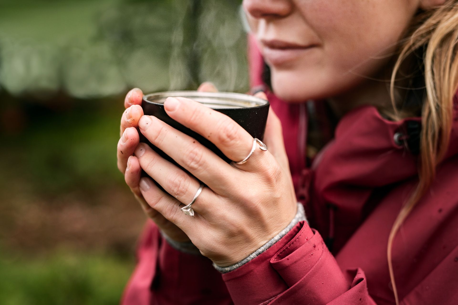 A woman in the woods is holding up a steaming cup of coffee.