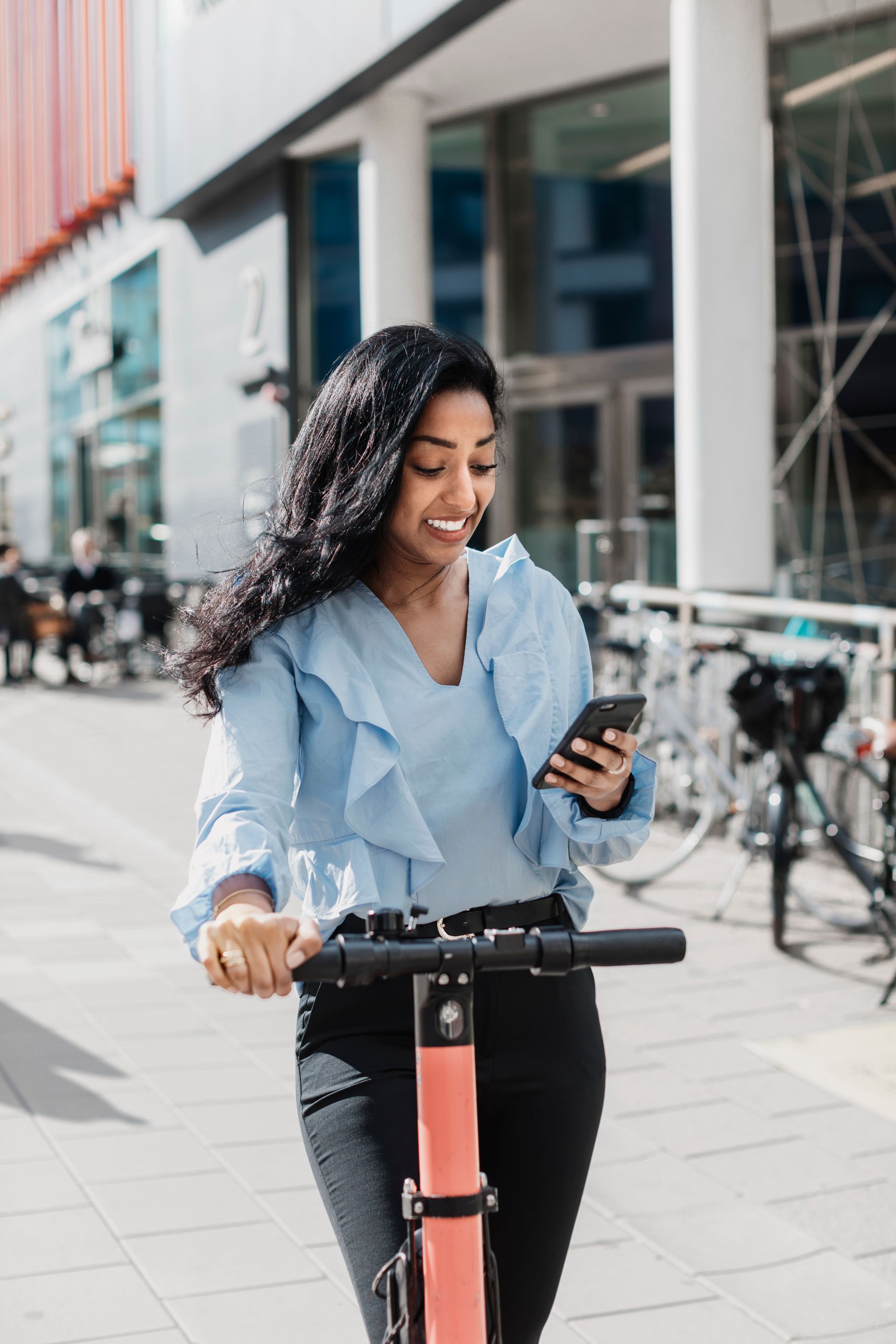 A woman is standing on an electric scooter outside, looking down at her phone.