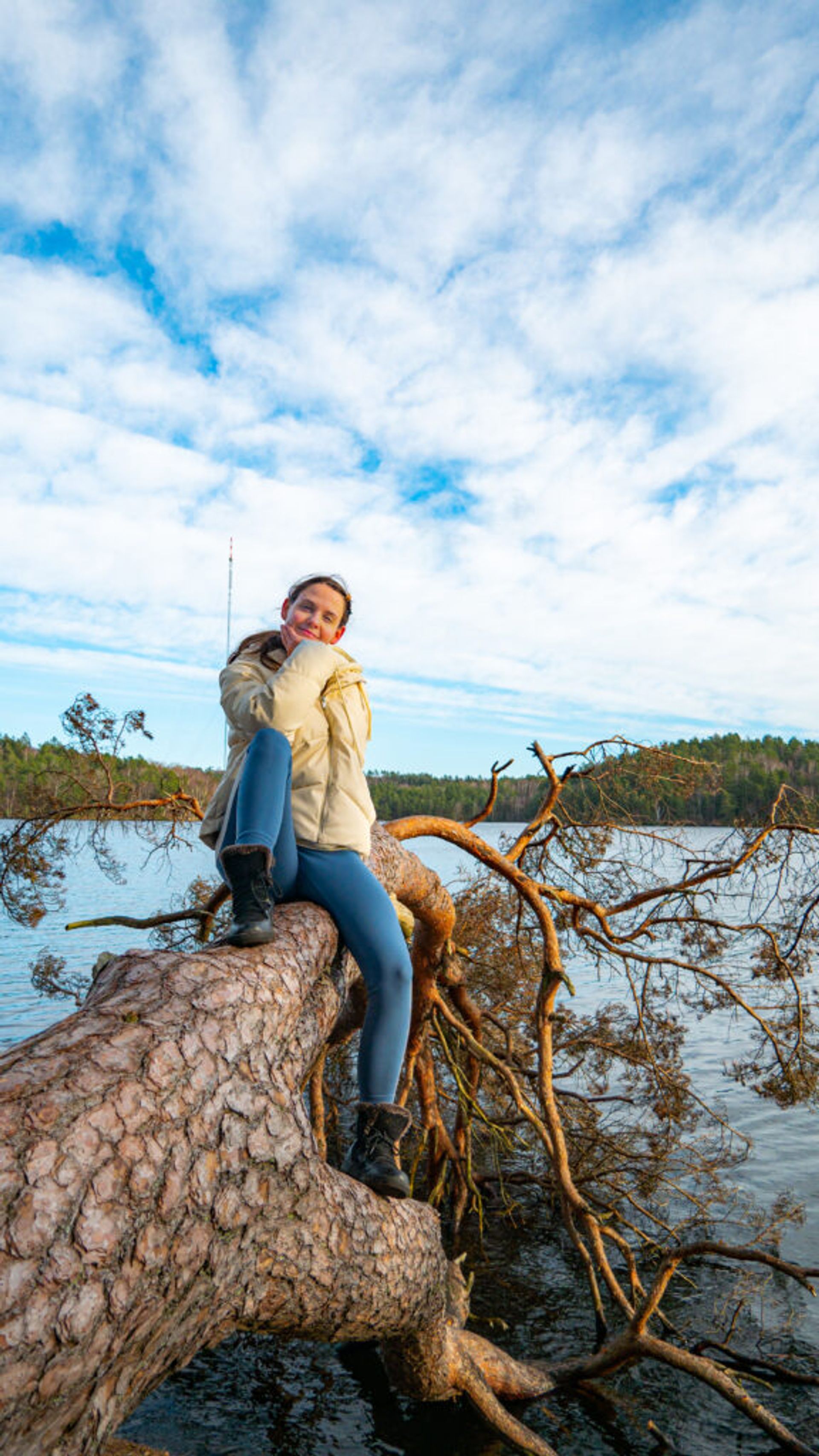 A woman poses on a fallen tree by a lake.