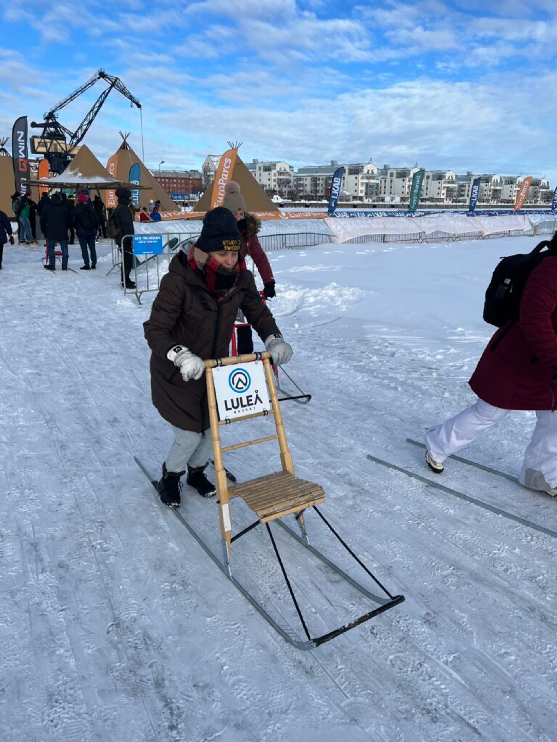 A woman kick-sledding on ice, with people in the background.