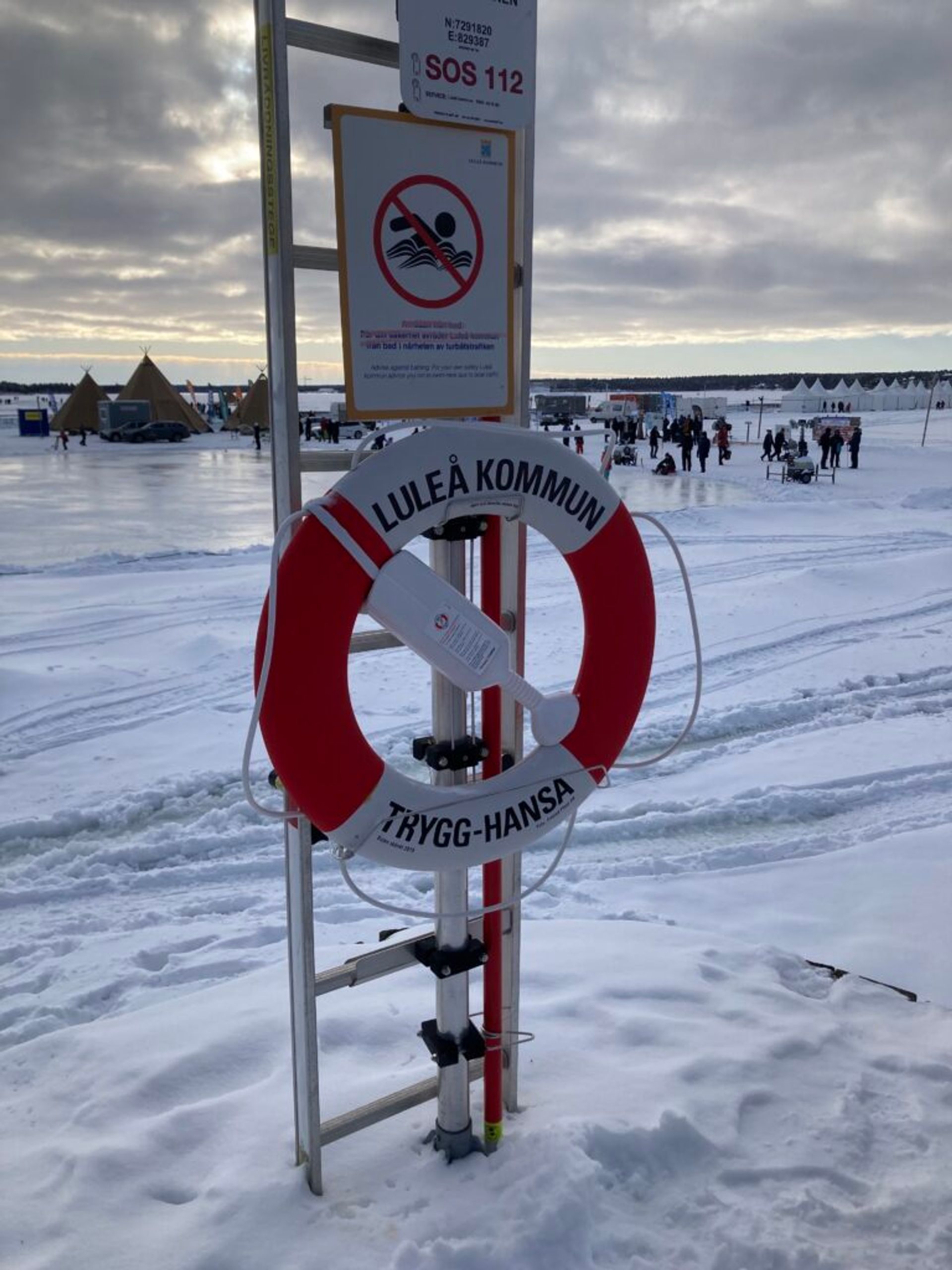 A lifebuoy attached to a safety station on a snowy lakeside with people and tents in the background.