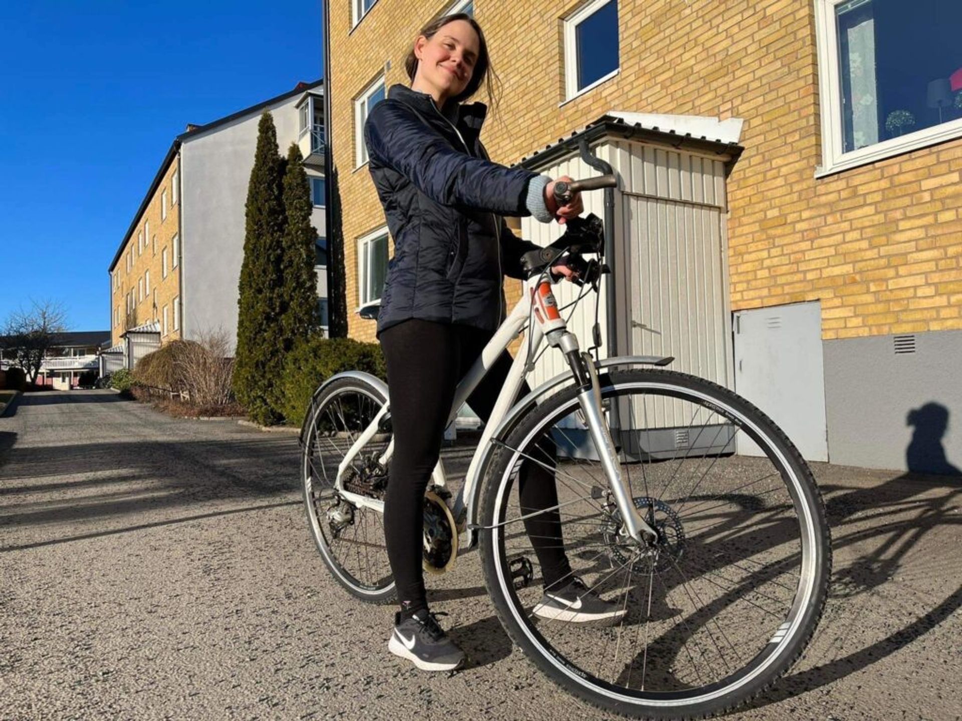 A woman posing happily with a bike beside a building.