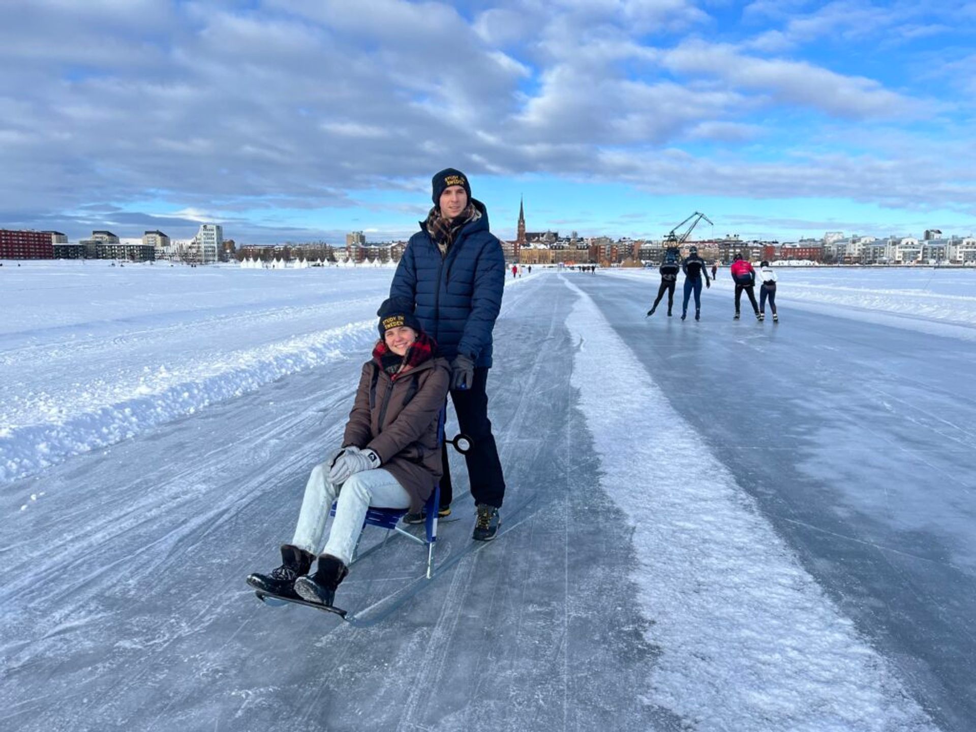 Two people, one seated on a kick-sled and another standing behind, on a vast outdoor ice rink with a cityscape in the background.