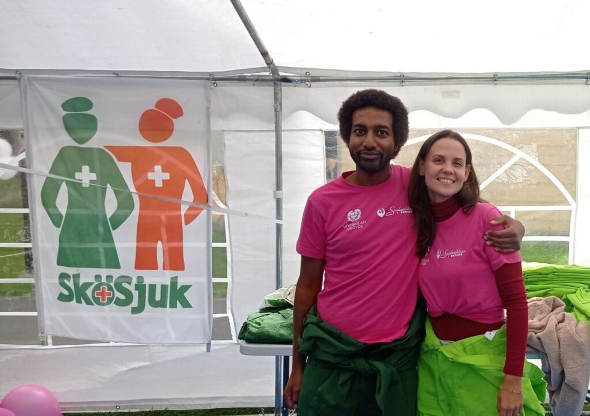 A man with his arm around a woman, both wearing pink T-shirts, stand under a tent at a campus event.