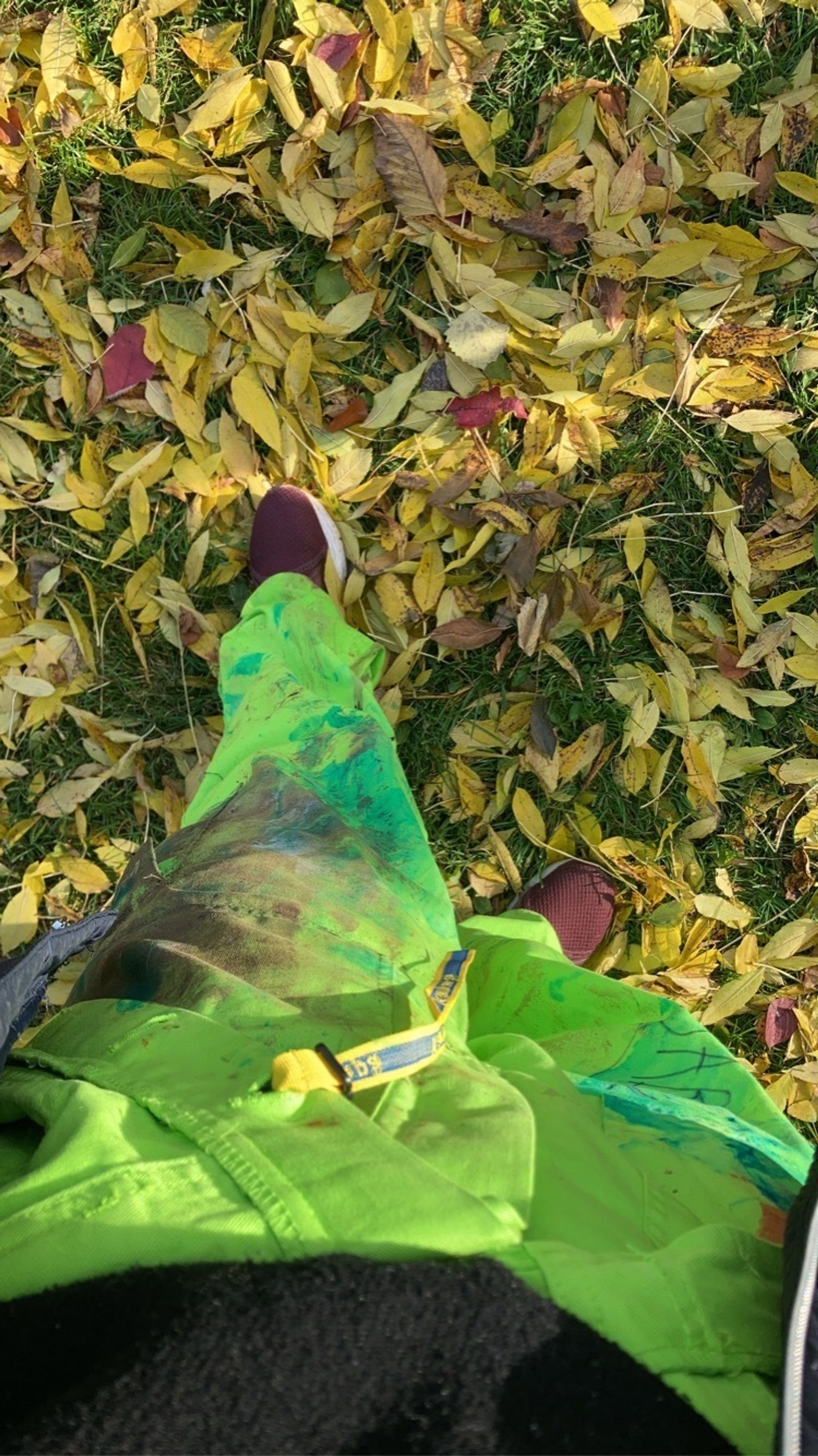 The lower half of a person wearing green overalls walks amidst fallen leaves.