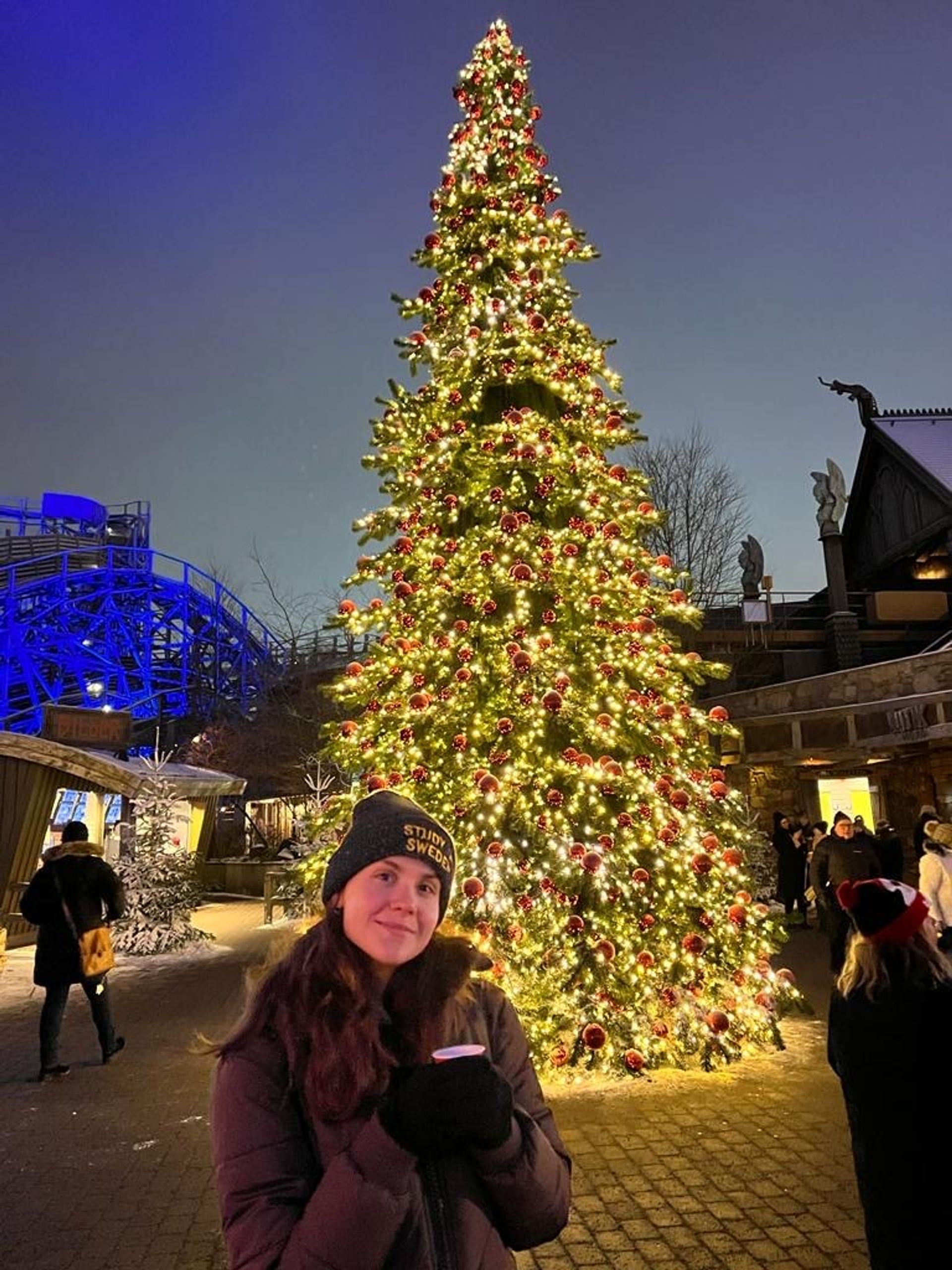 A woman stands in front of a beautifully lit Christmas tree, adorned with festive decorations, at a theme park.