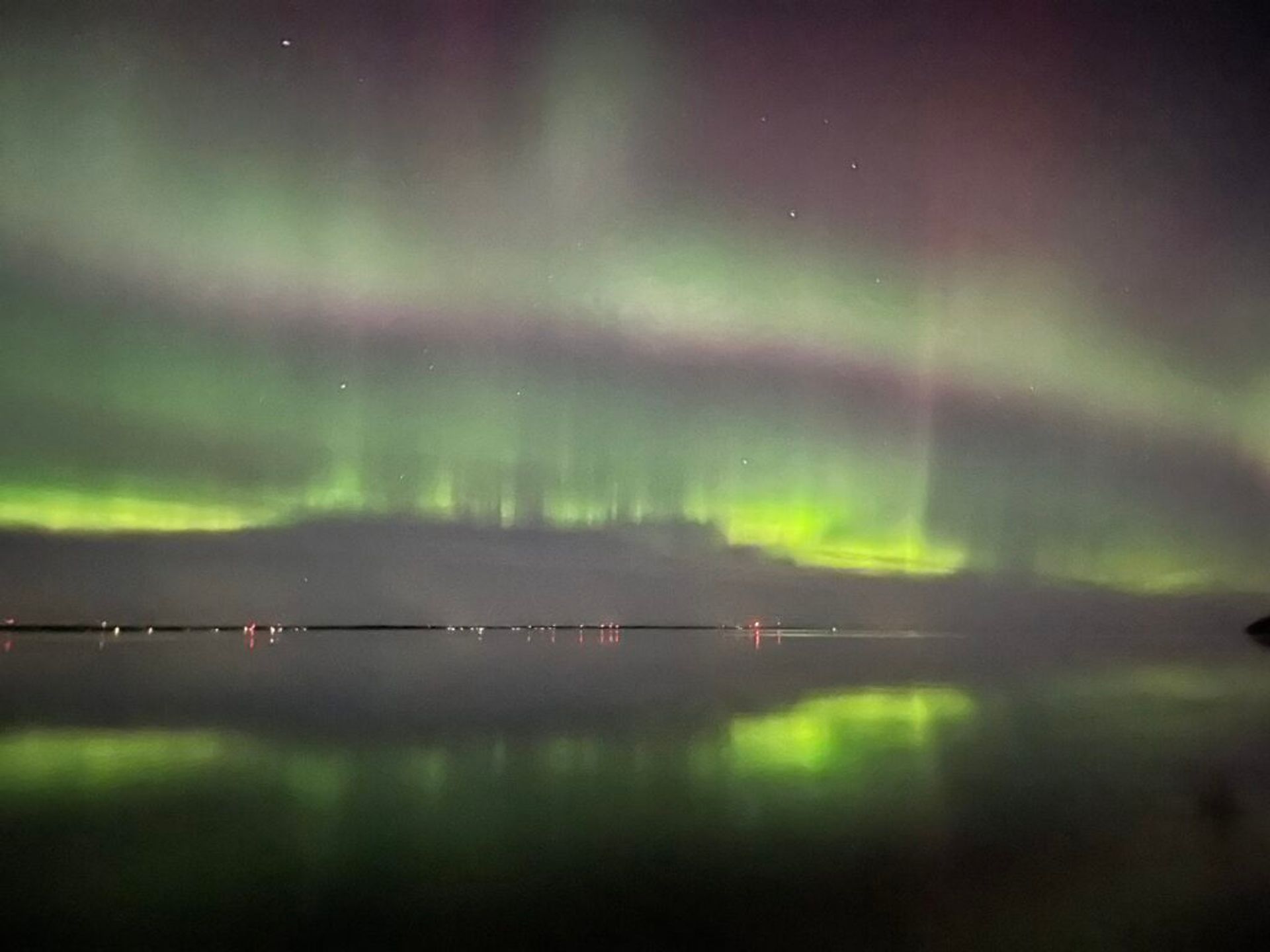 The Northern Lights cast a green glow over a tranquil landscape, mirrored in the still waters below.