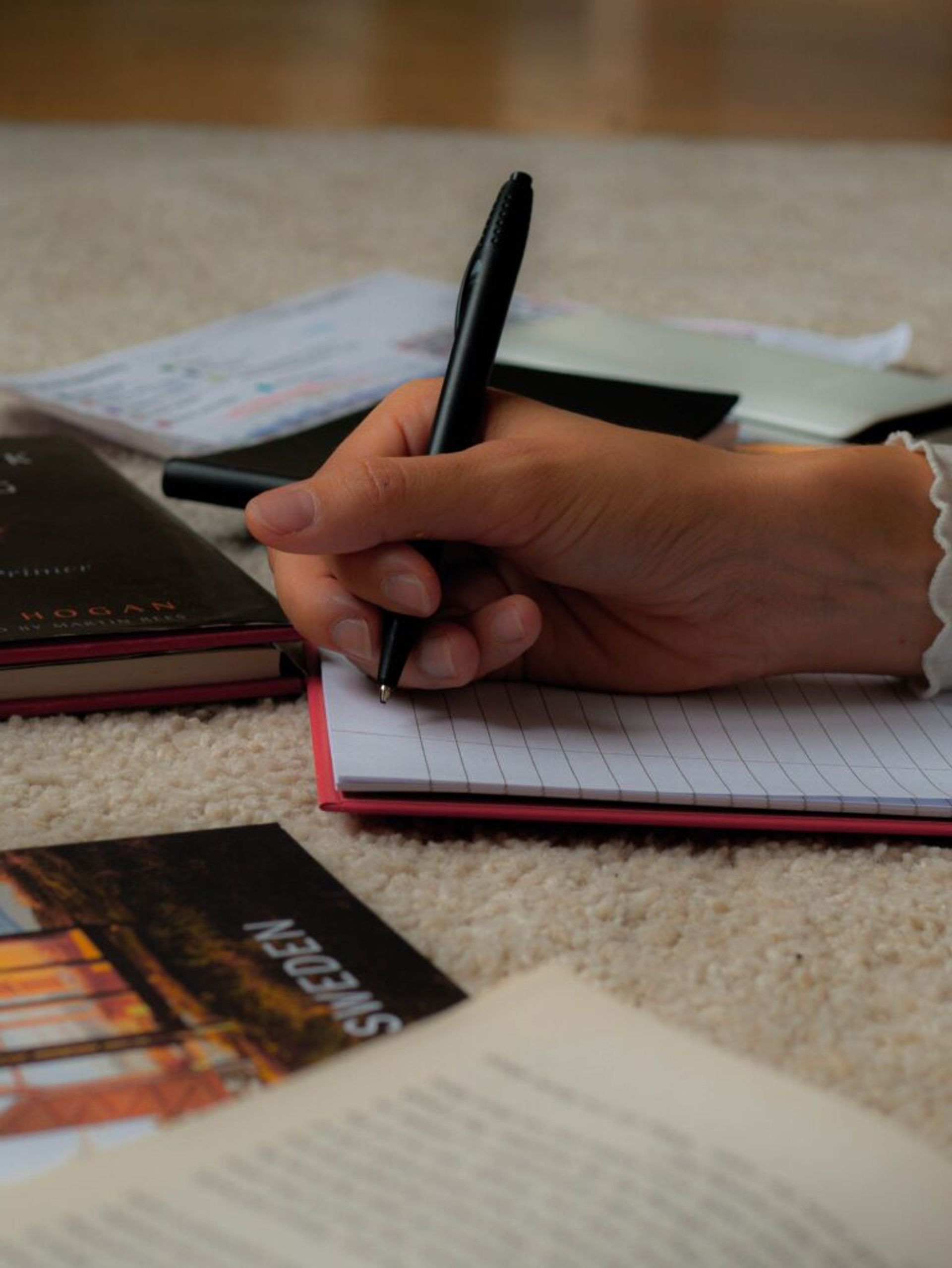 A hand holds a pen poised over a lined notebook, surrounded by study materials.