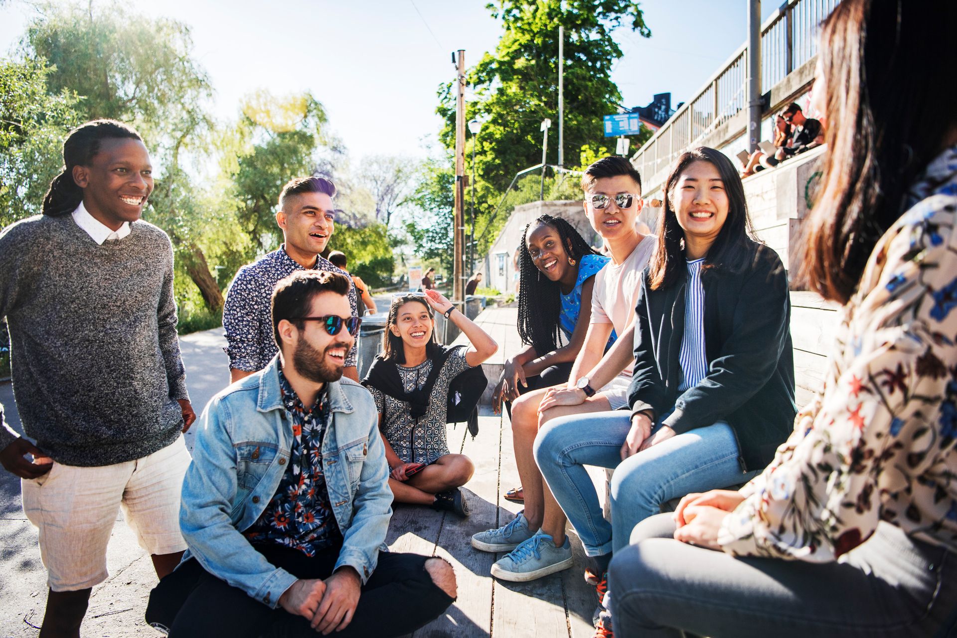 A group of diverse friends are gathered outdoors, enjoying each other's company in a sunlit, casual urban setting, engaging in conversation with expressions of laughter and happiness.