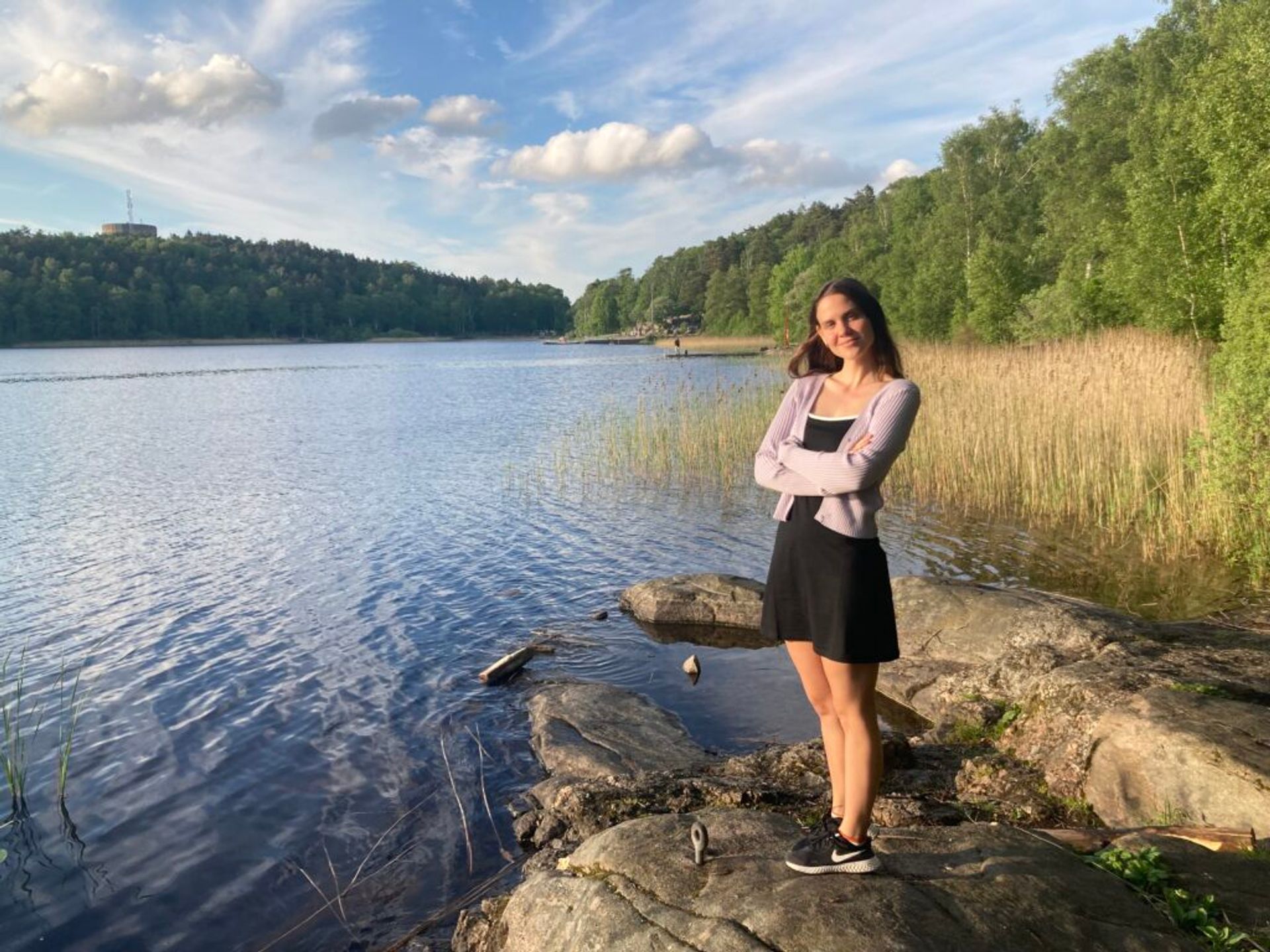 A woman with a contented smile stands by a lake, arms crossed, in a serene natural setting with calm waters, reeds, and a forested hill under a soft evening sky.
