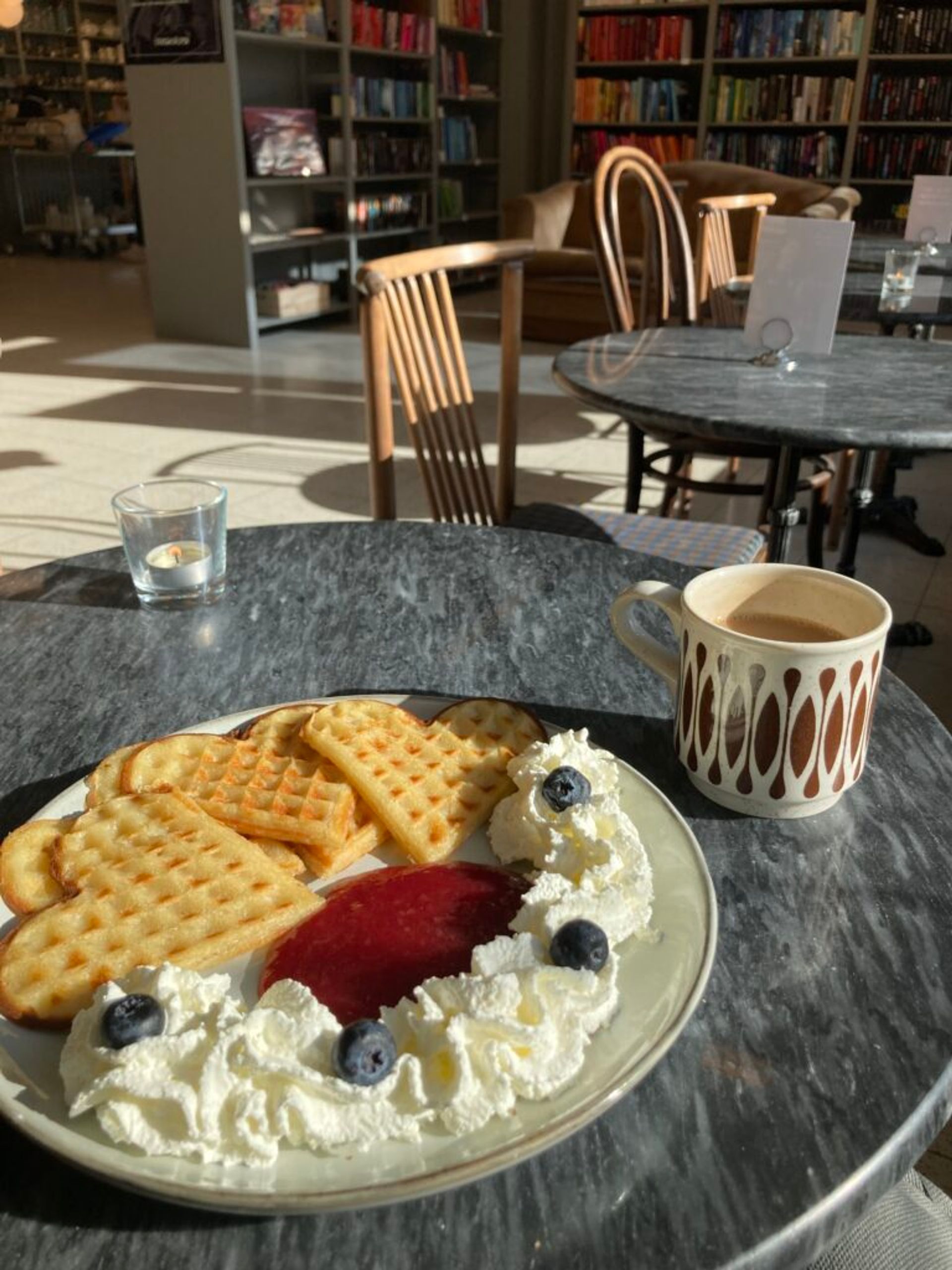 Heart-shaped waffles with whipped cream, blueberries, and jam, accompanied by a cup of coffee. The scene is set in a cozy indoor café.