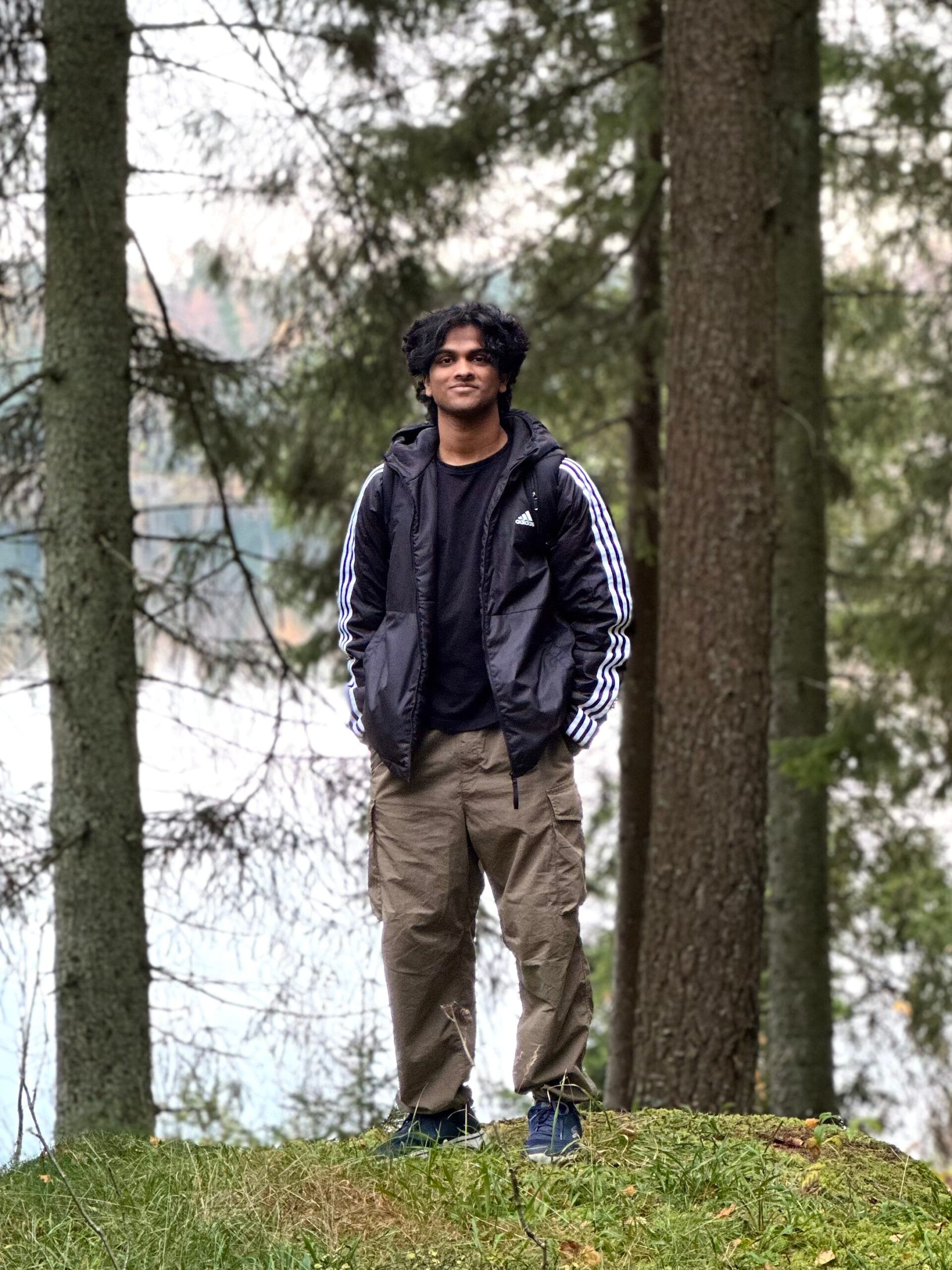 A boy standing in the middle of an opening in the forest, with a lake behind him