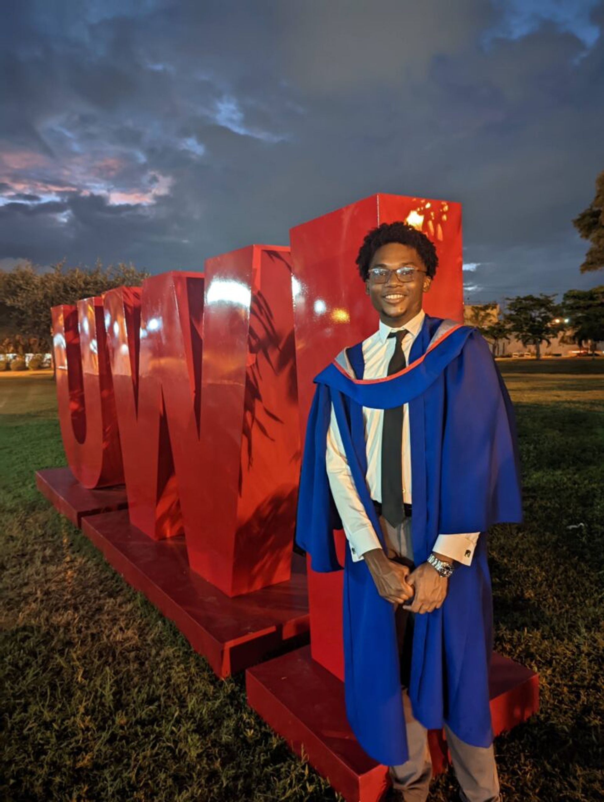 DA Joshua standing in front of the University of the West Indies (UWI) sign in his graduation gown