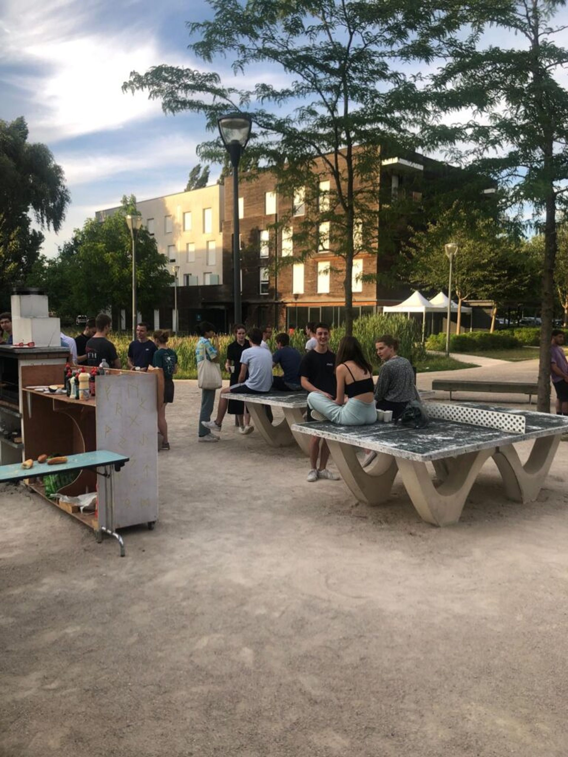 Photo of Chris' friends at a park in his city. Many people are sitting and talking on concrete table tennis sets in the afternoon sun. 