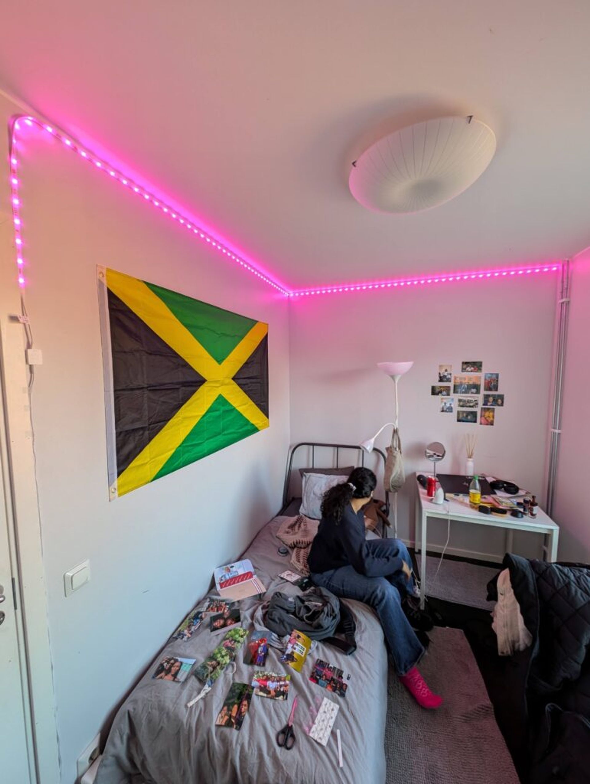 A picture of a bedroom with a Jamaican flag on the wall and a strip of lights along the top corner of the room illuminated in pink 