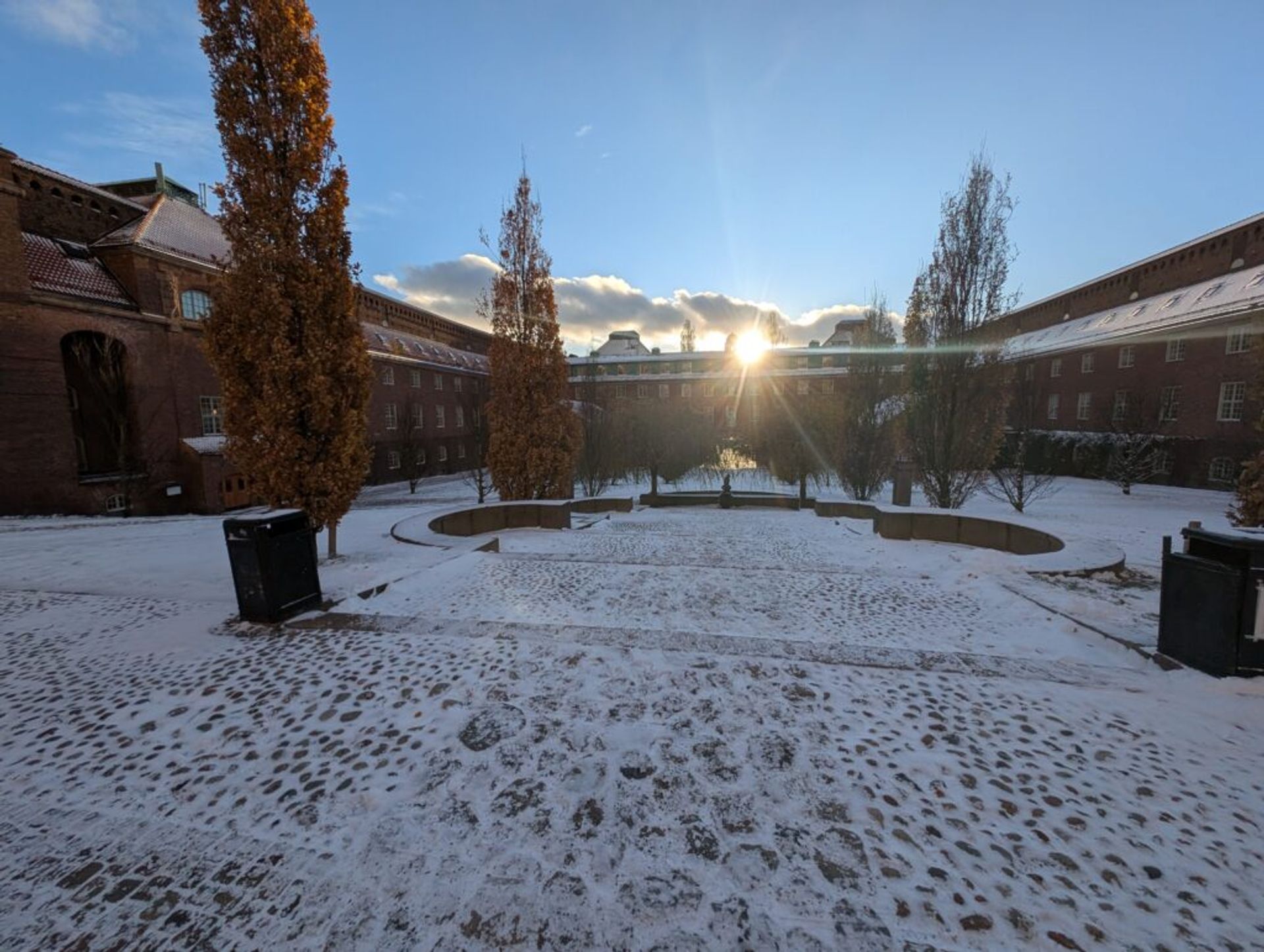 A picture of the snow covered courtyard at KTH Royal Institute of Technology. There is a cobblestone courtyard surrounded by brick buildings with the afternoon sun setting above the building to the west.