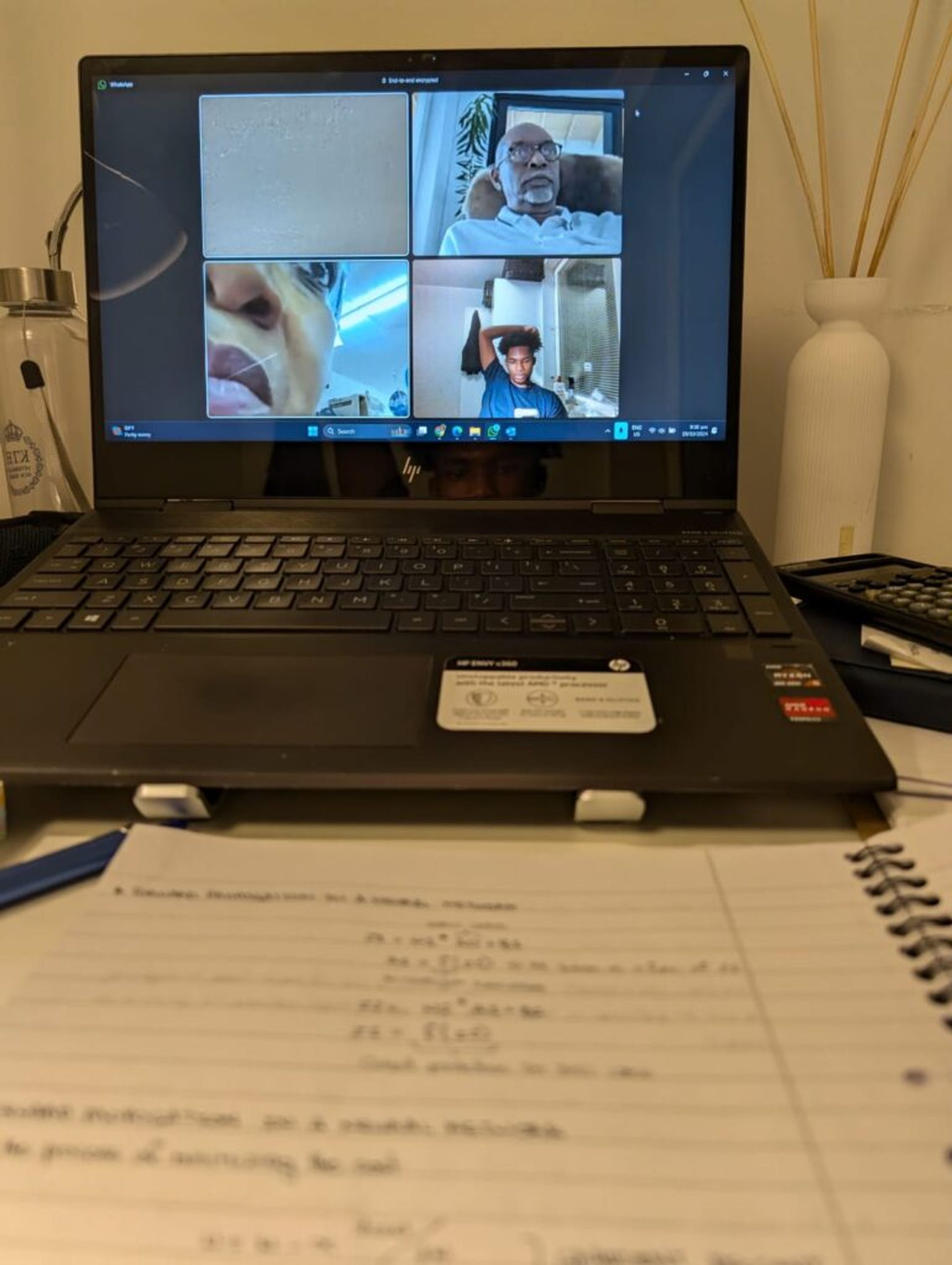 Picture of a laptop and a book on a desk. On the laptop screen is Joshua's family in a Google Meets call with four participants, each doing their own activities. 