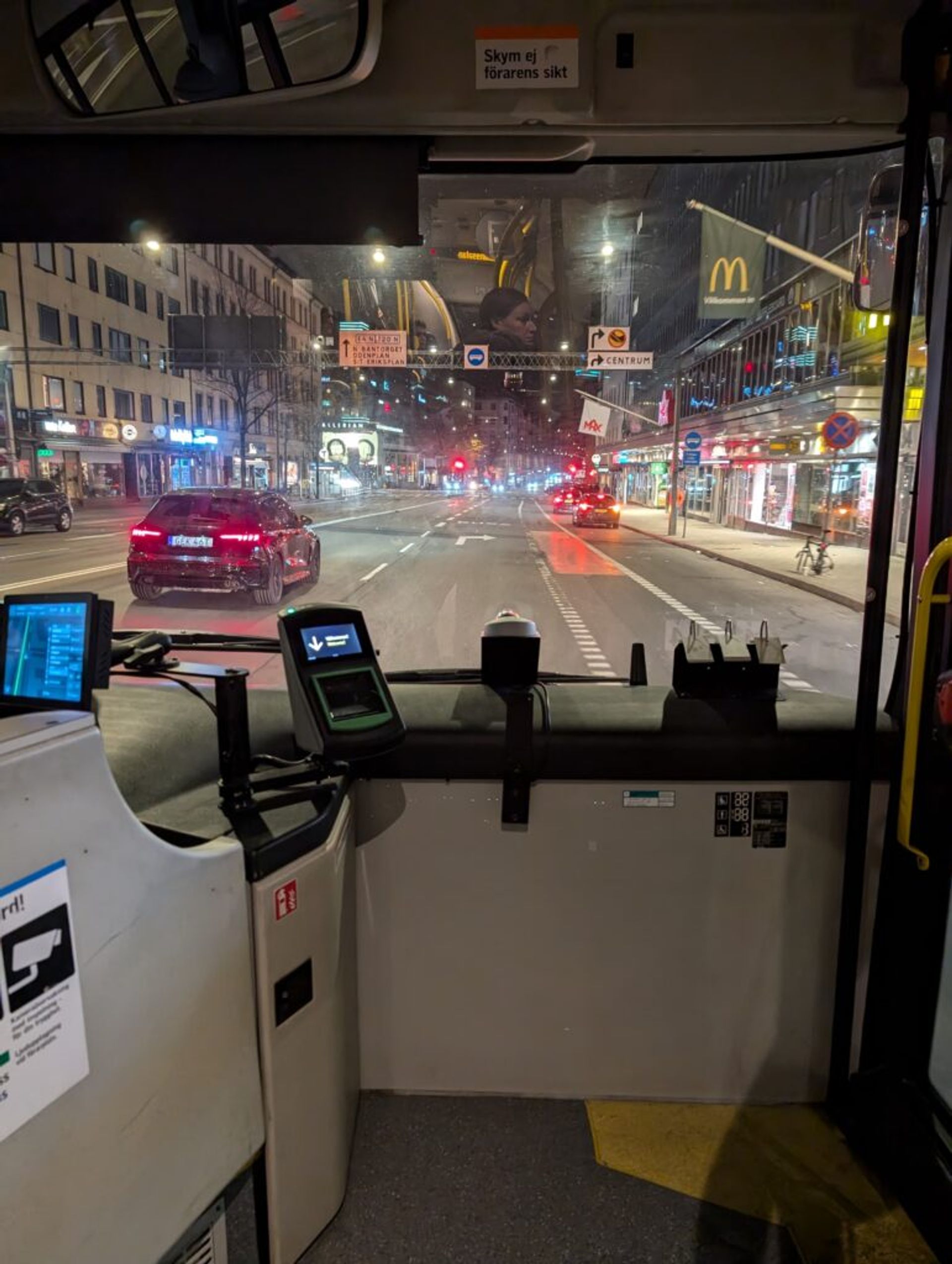 A photo looking out onto a street in the city through the windscreen of a bus. There are buildings on either side of the road and the street is not very busy. 