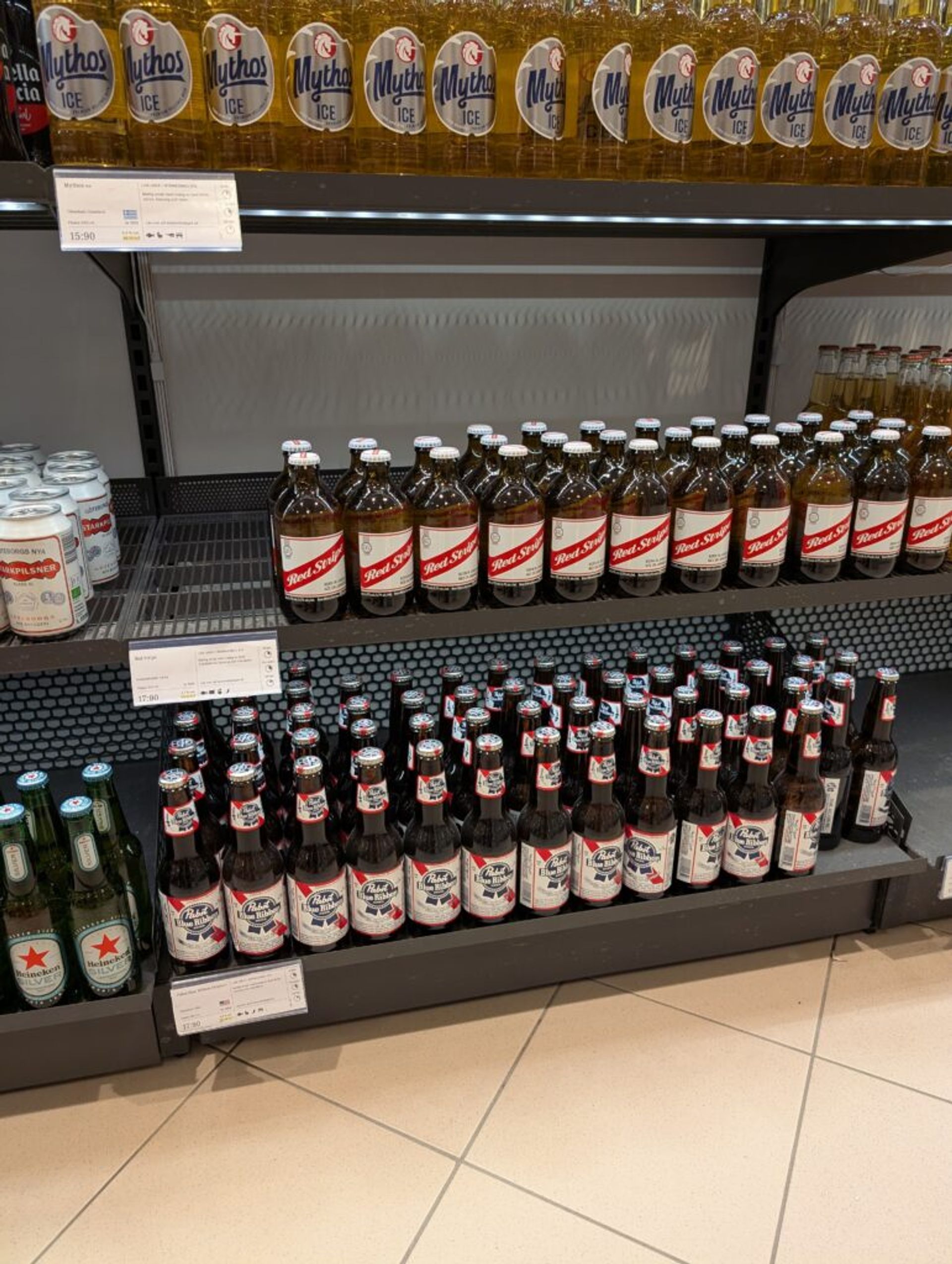 A picture of beer on a shelf in Systembolaget in Sweden. There are different types of beer including Mythos ICE, Red Stripe and others.