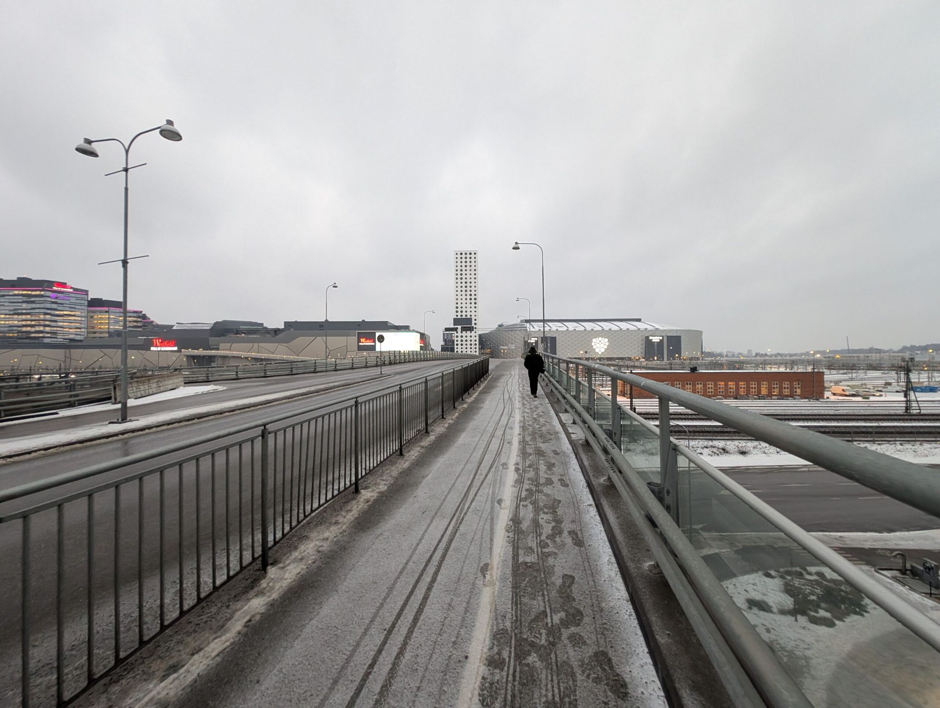 A snow covered bridge in Solna, Sweden. This picture is taken from the point of view of someone crossing the bridge. There is one person distantly ahead and some big buildings and a grey sky in the background. 