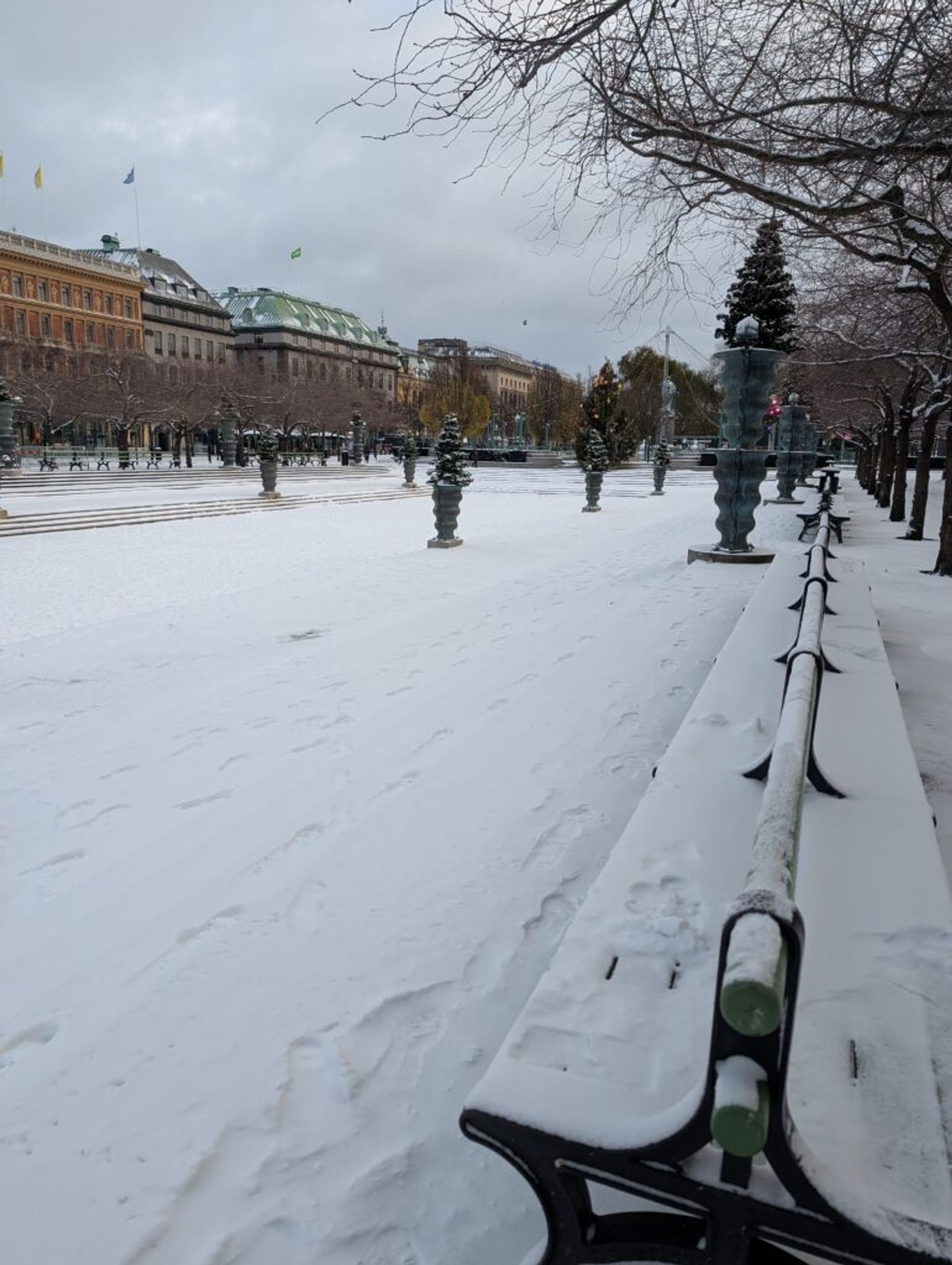 A photo looking into a snow covered concrete park. There is fluffy snow on the ground and on the rooves of the buildings in the background.