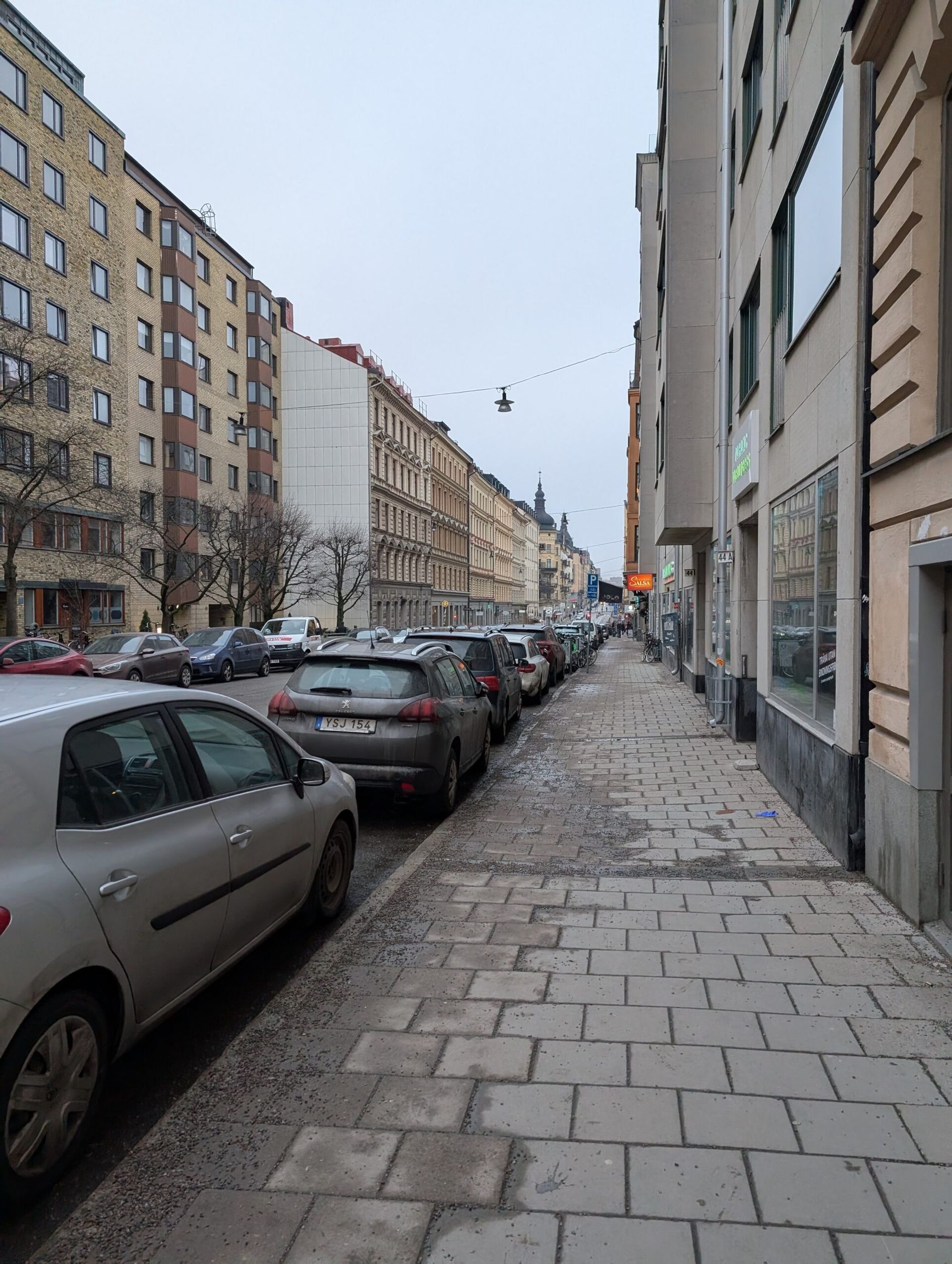 Looking down the street in Stockholm. The picture is taken from the sidewalk with cars against the curb and buildings in different shades of yellow on both sides of the street