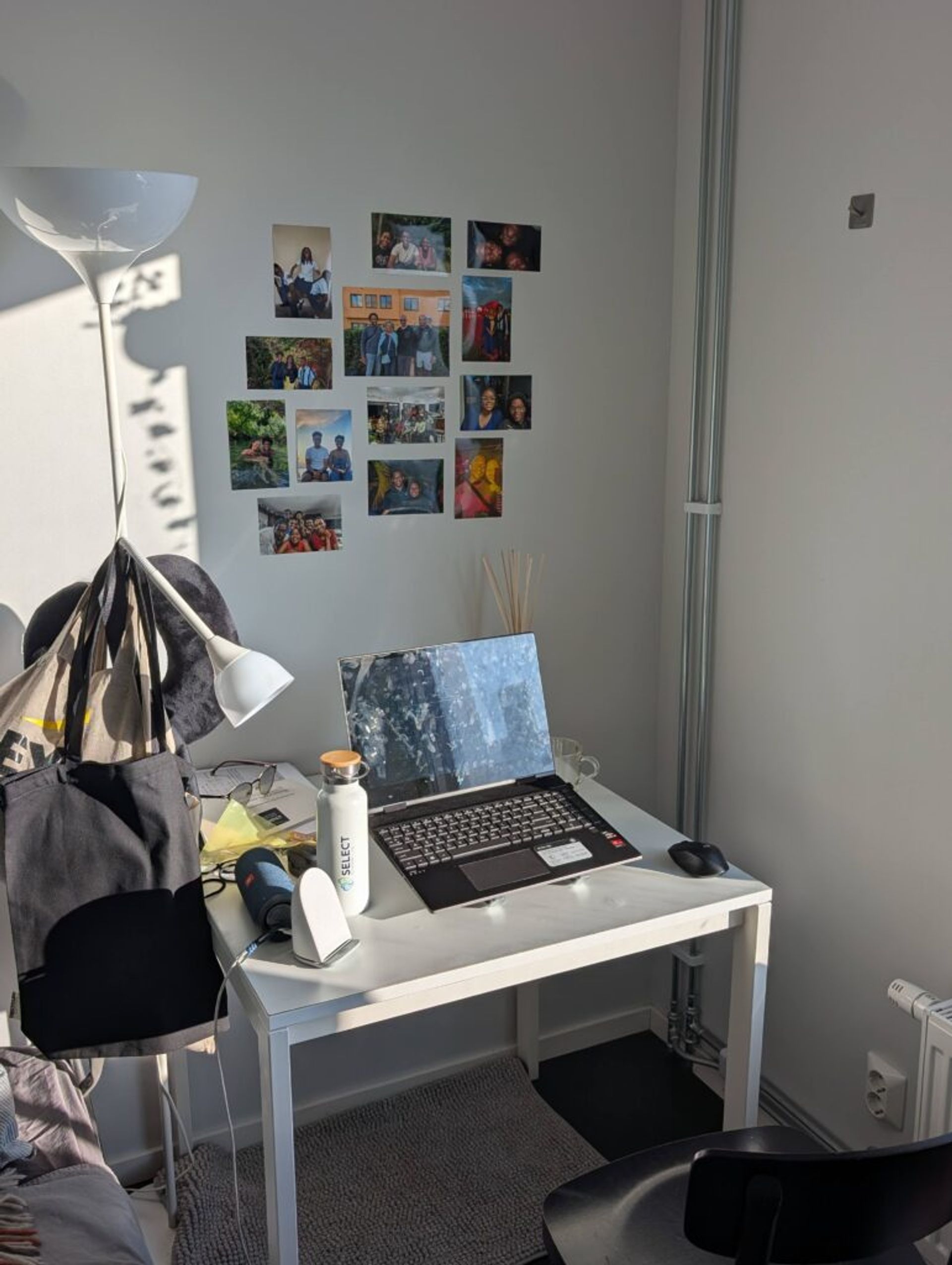 Picture of the sun shining into Joshua's bedroom desk with pictures of his family and friends above the desk. A laptop, speaker, books and other items are on the white table.