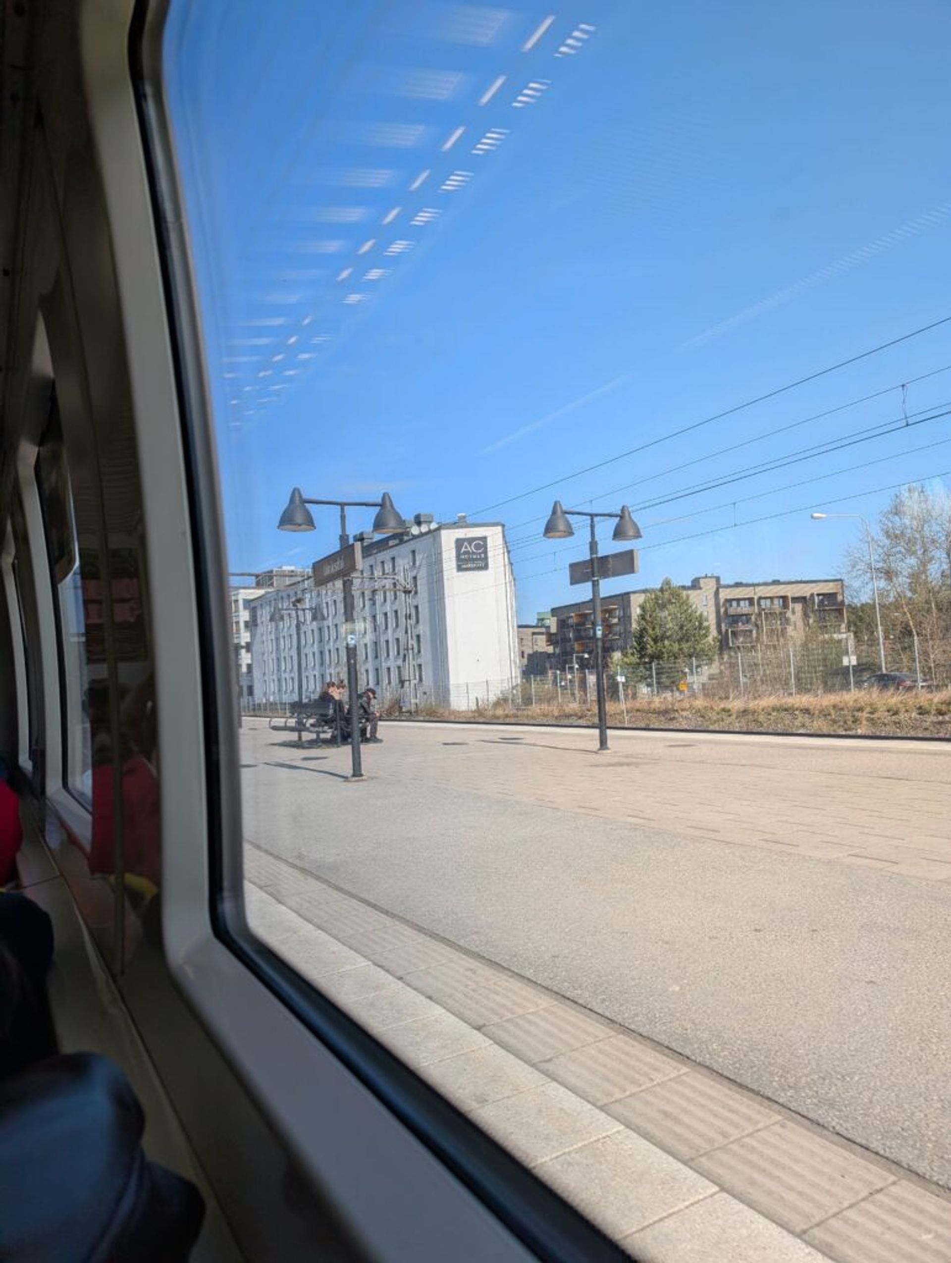View out the window of a train. Through the window is the platform with seating and buildings in the background.