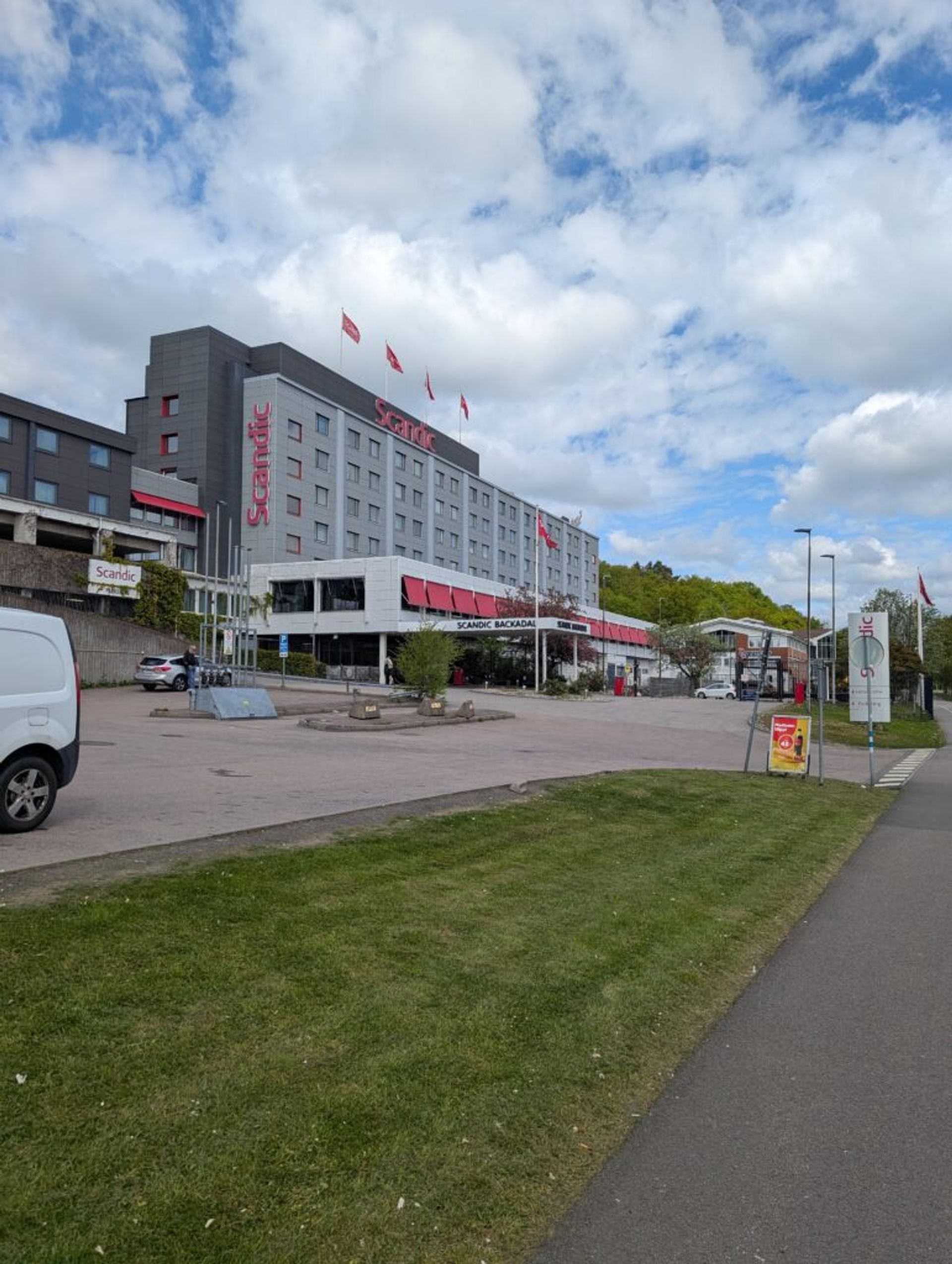 A picture of a Scandic Hotel. The building is grey and brown with the red Scandic logo and Scandic flags. The sky above the building is cloudy but blue in places.
