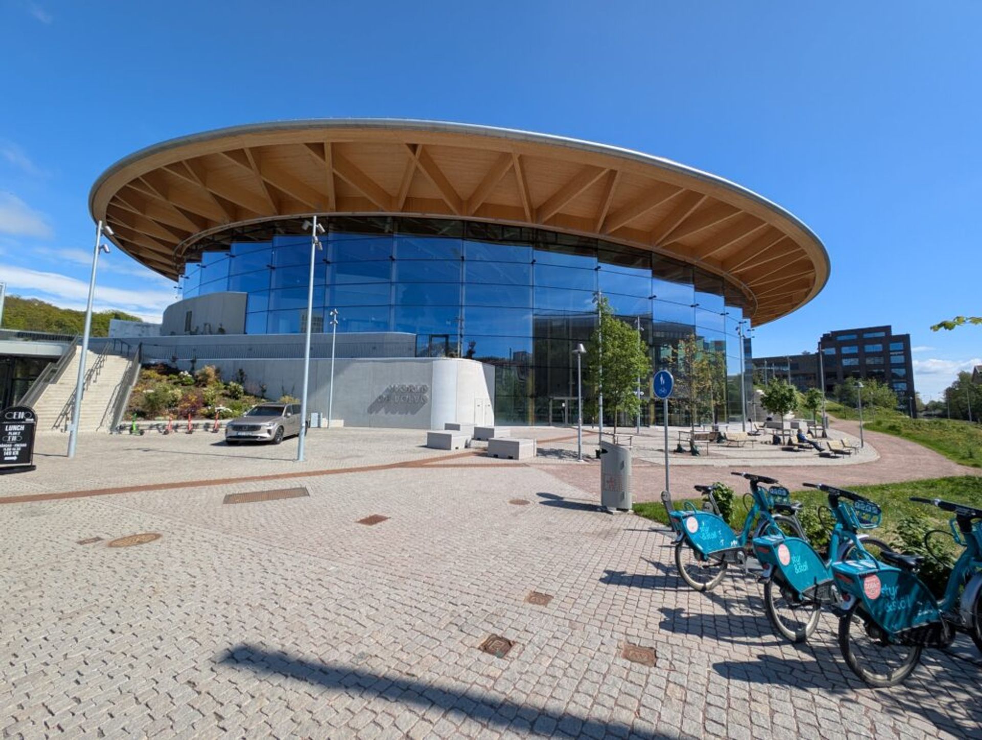 The 'World of Volvo' building. The building is cylindrical and covered with glass with a wooden overhang. The picture is taken with the paved stone walkway in the foreground and almost completely clear blue sky in the background.
