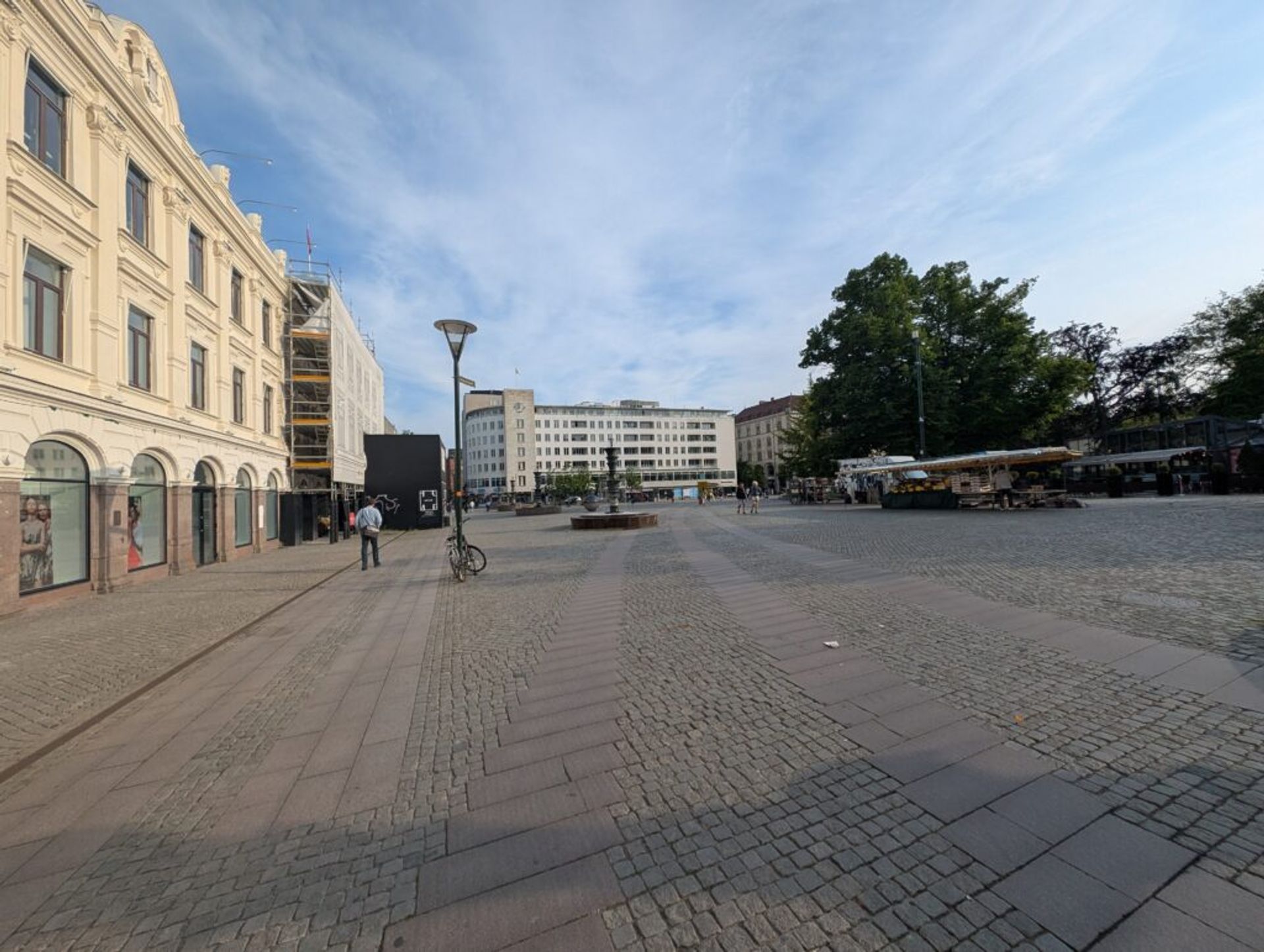 A picture of a town square in Malmö. The square is paved with brown paving stones and bordered by beige colored multi story buildings. The sky above the square is pale blue.