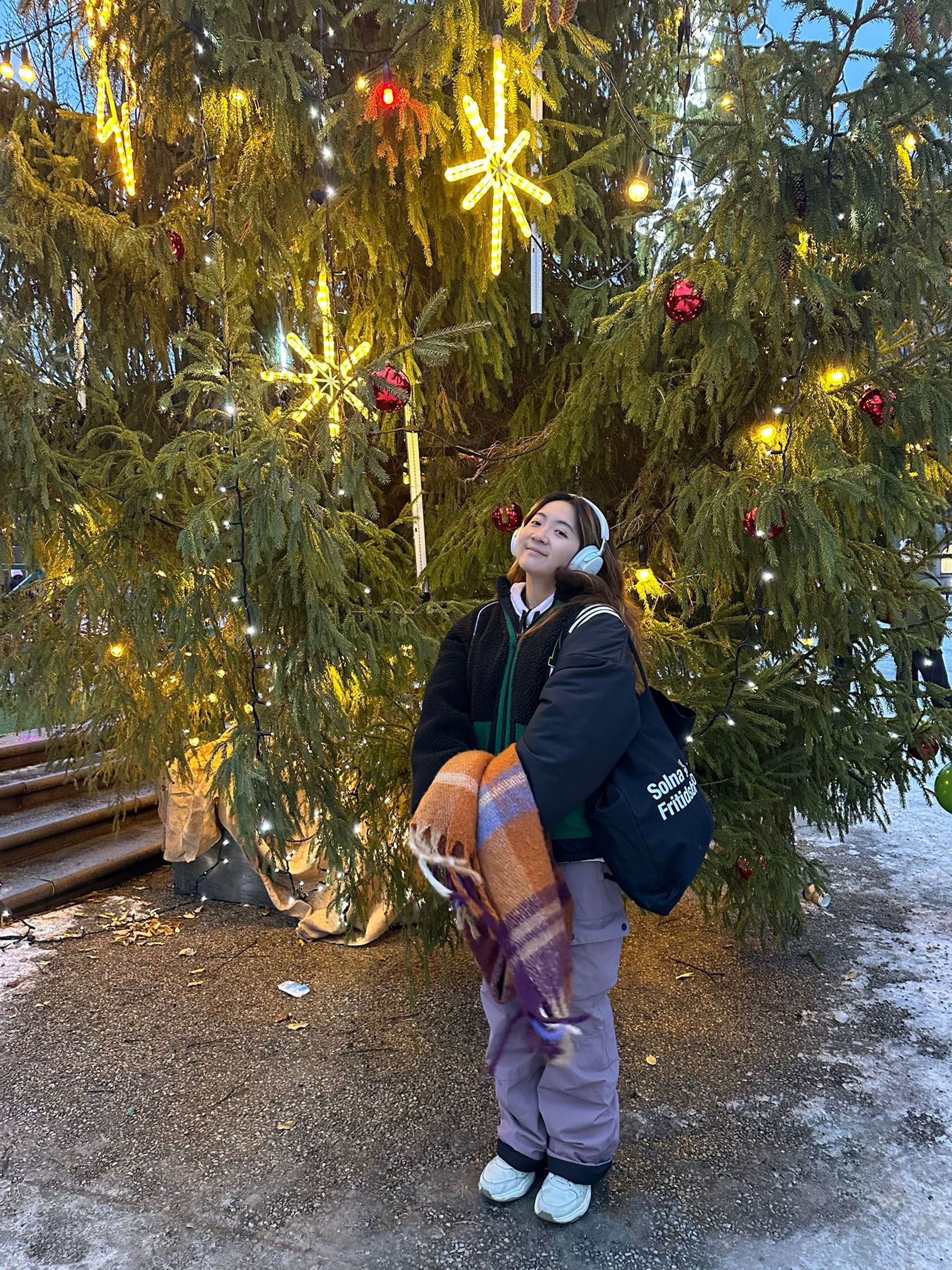 Yan-Lin posing in front of a tree decorated with pepper lights in the winter. She is in winter clothing, holding a scarf