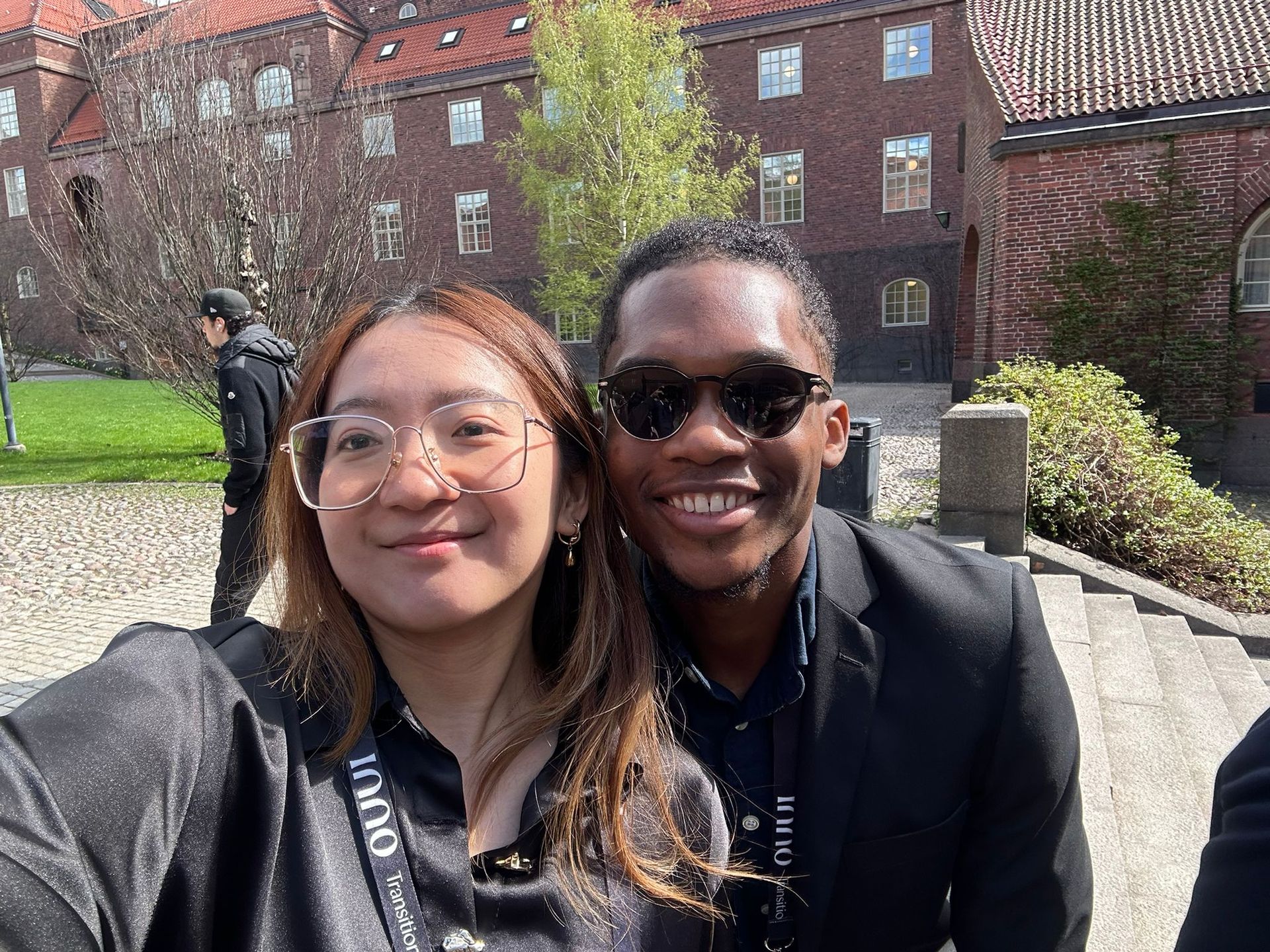 Joshua and Yan-Lin posing for a selfie. They are both in formal wear with lanyards and red brick buildings in the background.