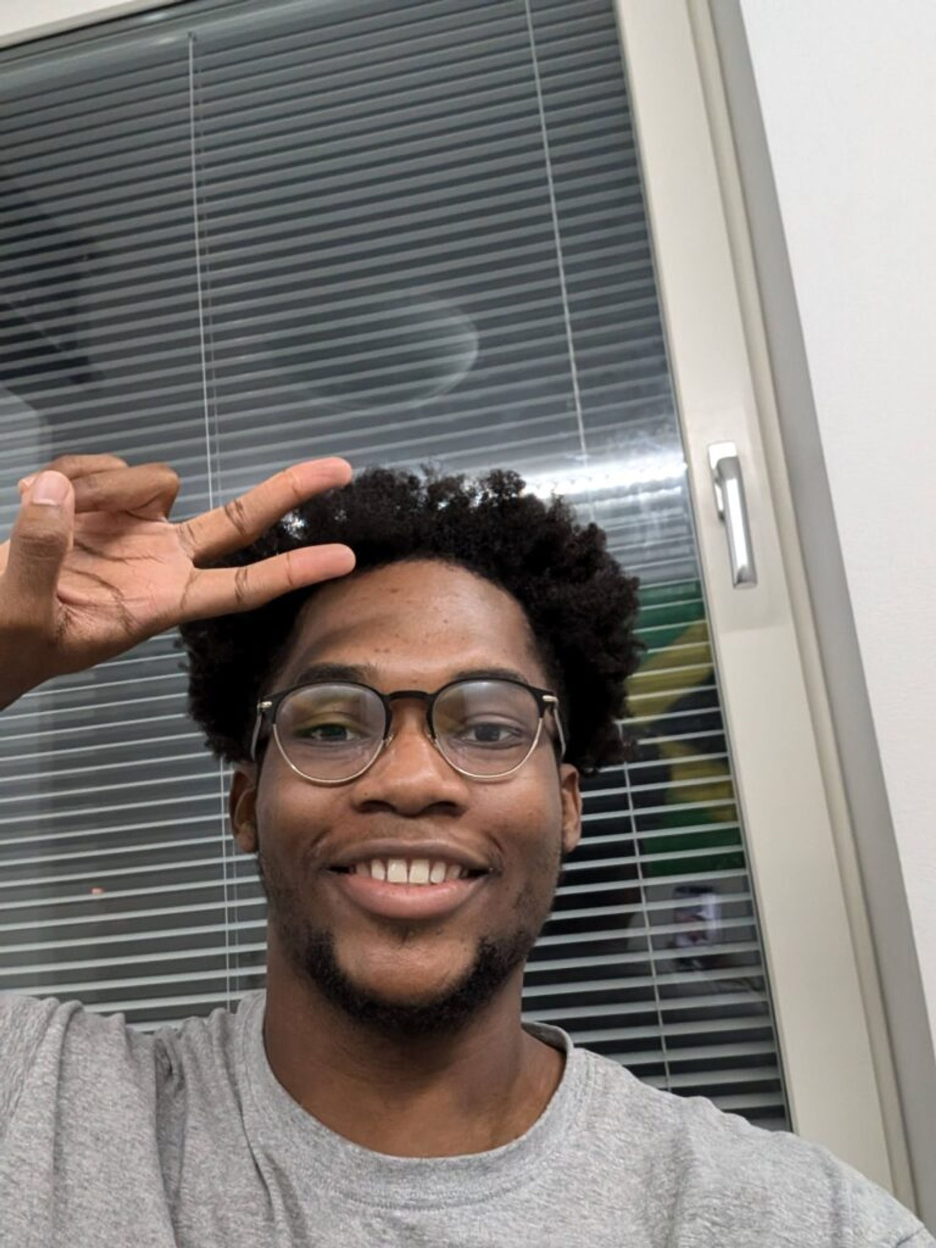 A photo of Joshua smiling in front of a window with blinds. He is posing with a peace sign.