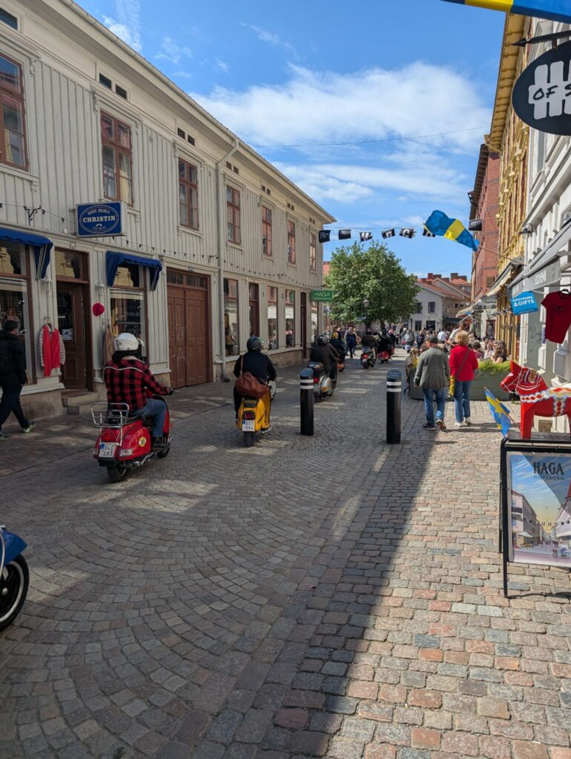 Picture of a cobblestone walkway in the old town in Gothenburg. There are people walking in the sun and Vespa scooters driving down the middle of the pathway. The pathway is lined with old buildings that have shops.