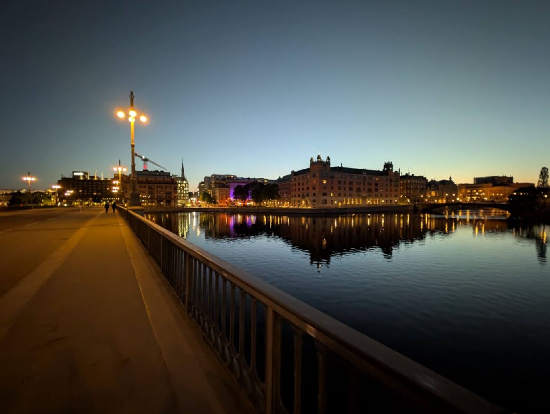A photo on the sidewalk of a bridge at night. There is water under the bridge and there are buildings on the edge of the water illuminated by amber lights.