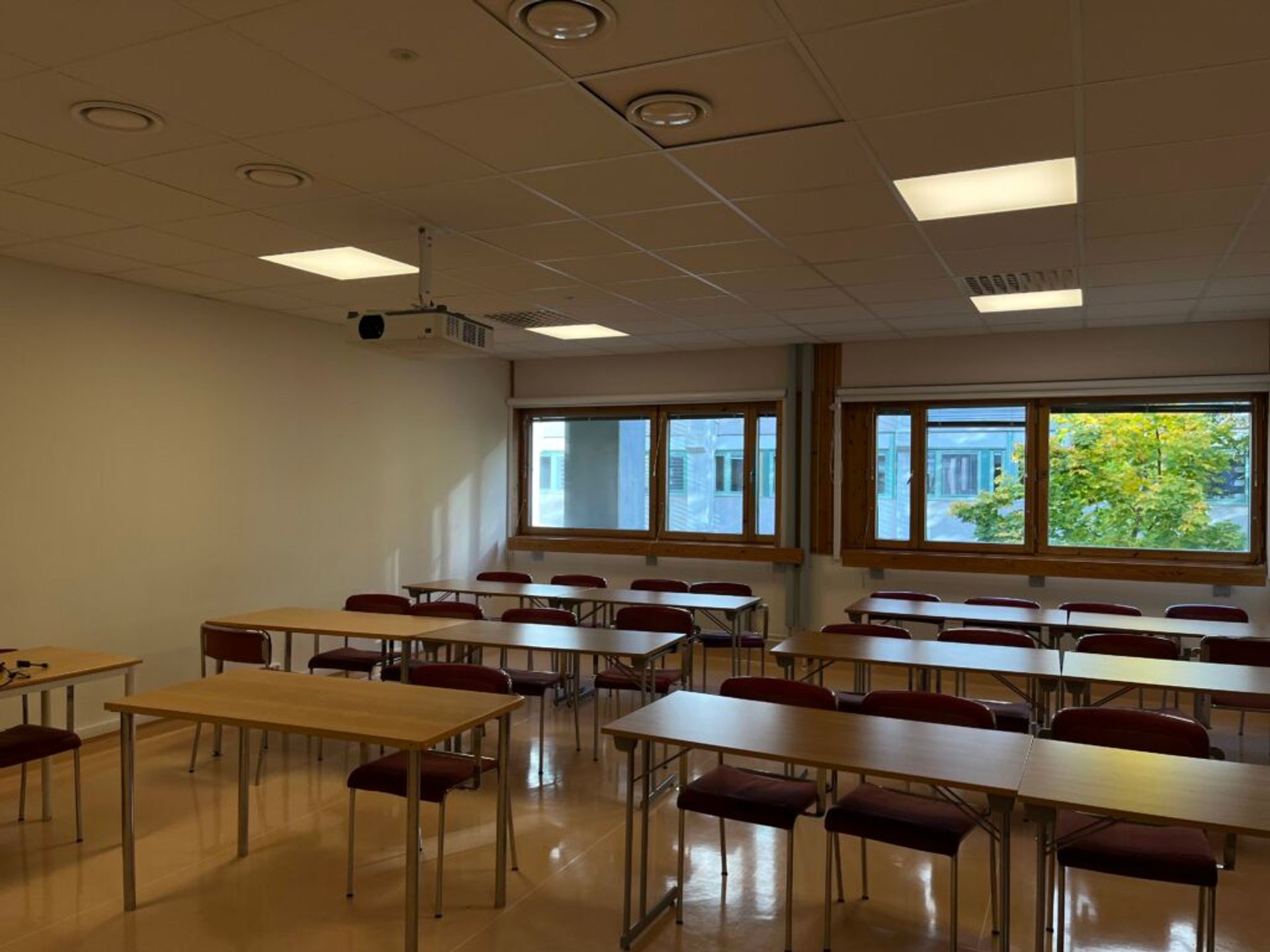 An empty classroom with approximately 24 seats. Wooden windows with a colourful tree outside is to be seen in the back.