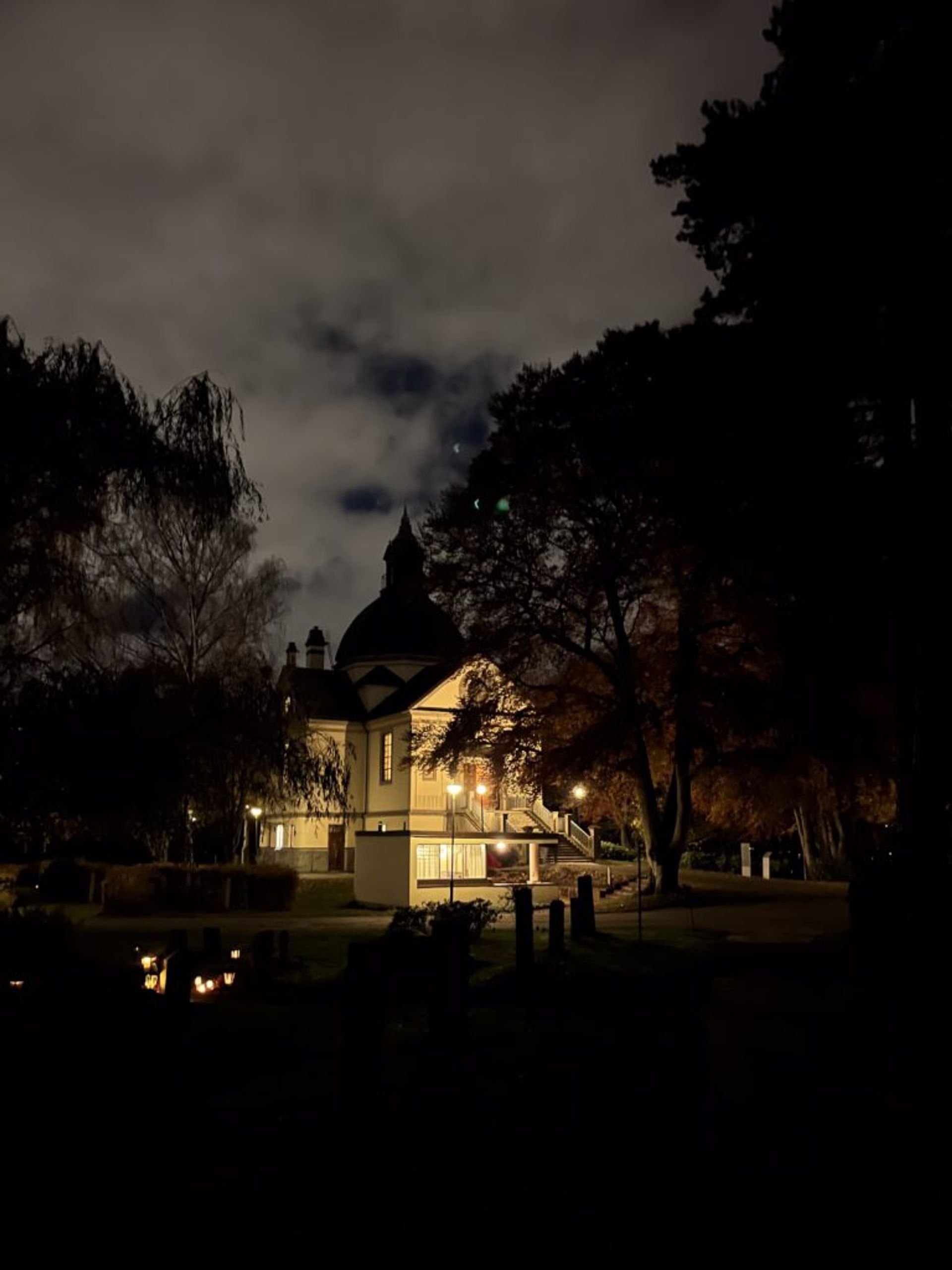 A lit, pale yellow chapel, partially covered by reddish trees.